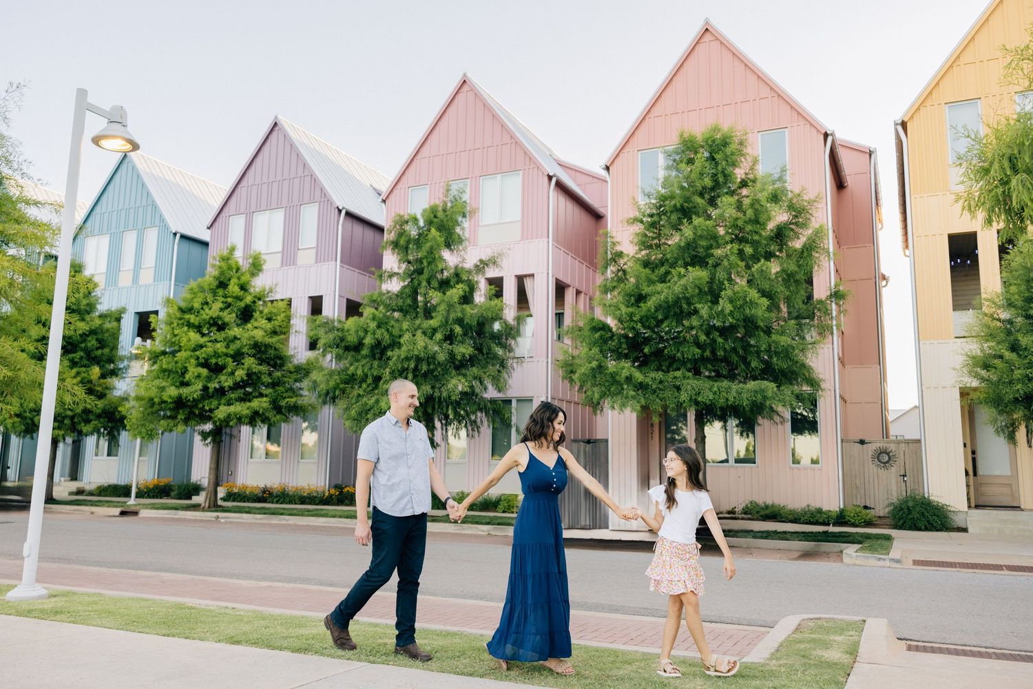 Three people walking past modern pastel-colored townhouses with peaked roofs and green trees along a residential street.