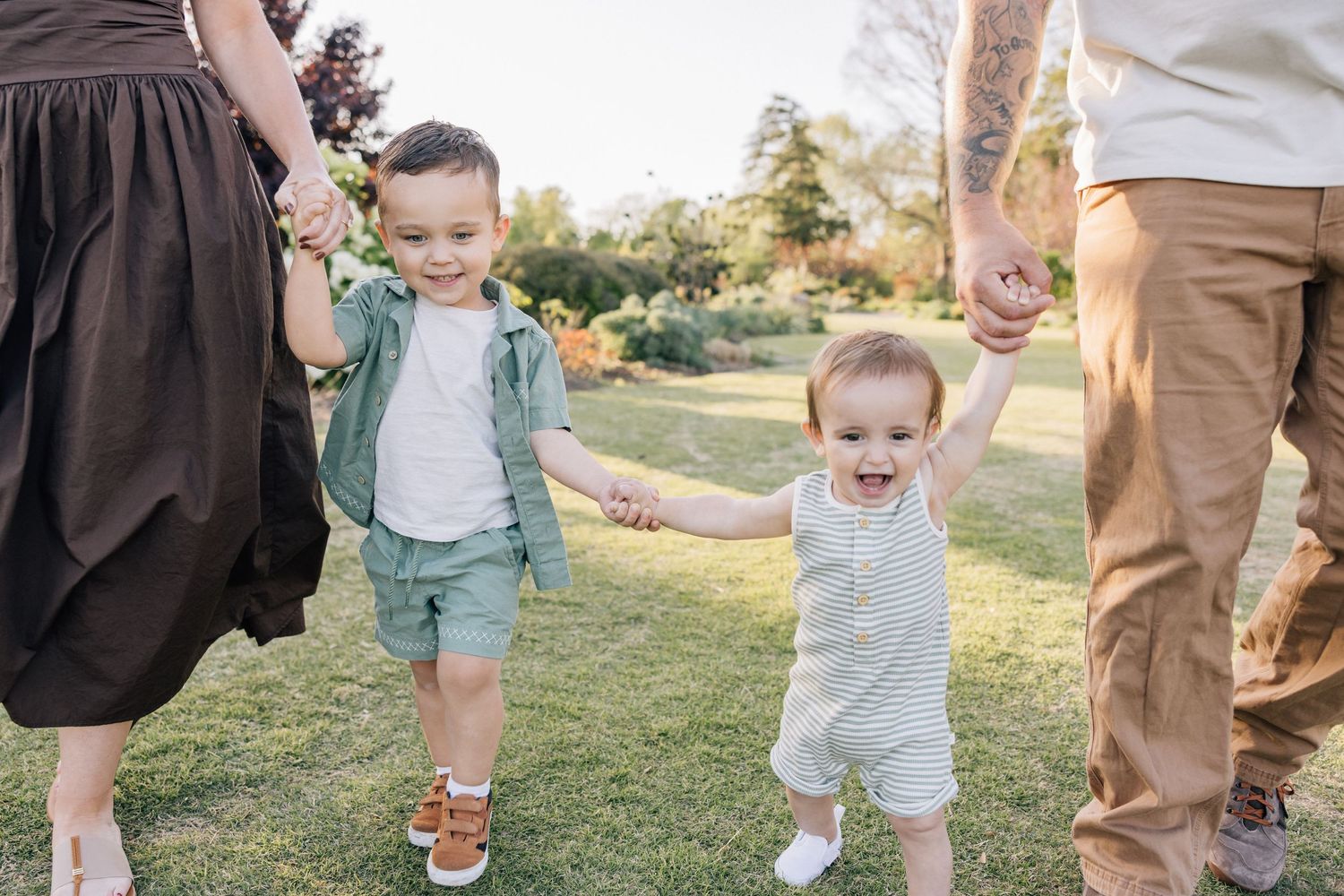 A toddler and a young child walk hand in hand with their parents in a sunny garden setting.