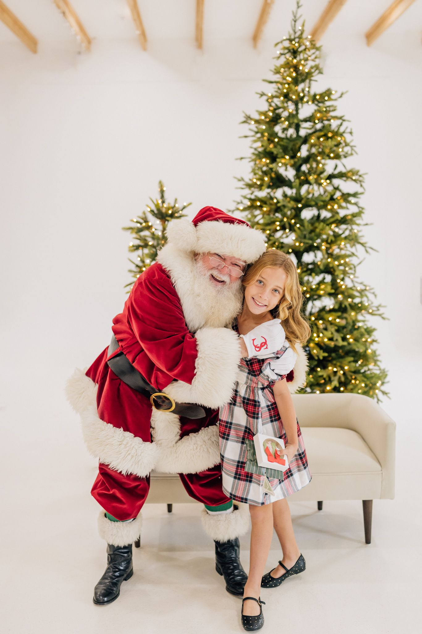 A cheerful girl in plaid dress hugs Santa Claus beside a glowing Christmas tree in a bright white studio.