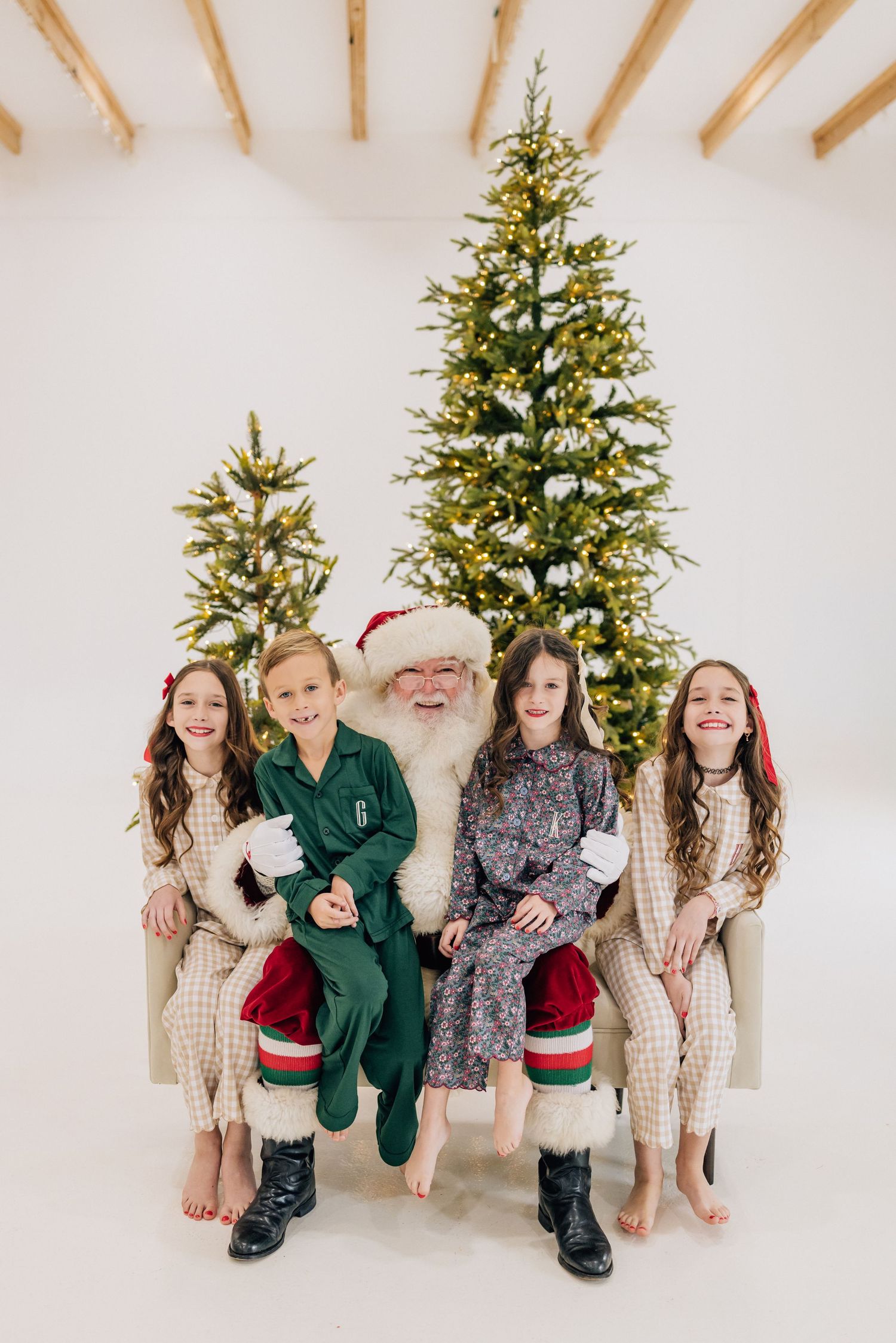 Four children sit with Santa Claus in front of lit Christmas trees in a bright white studio setting.