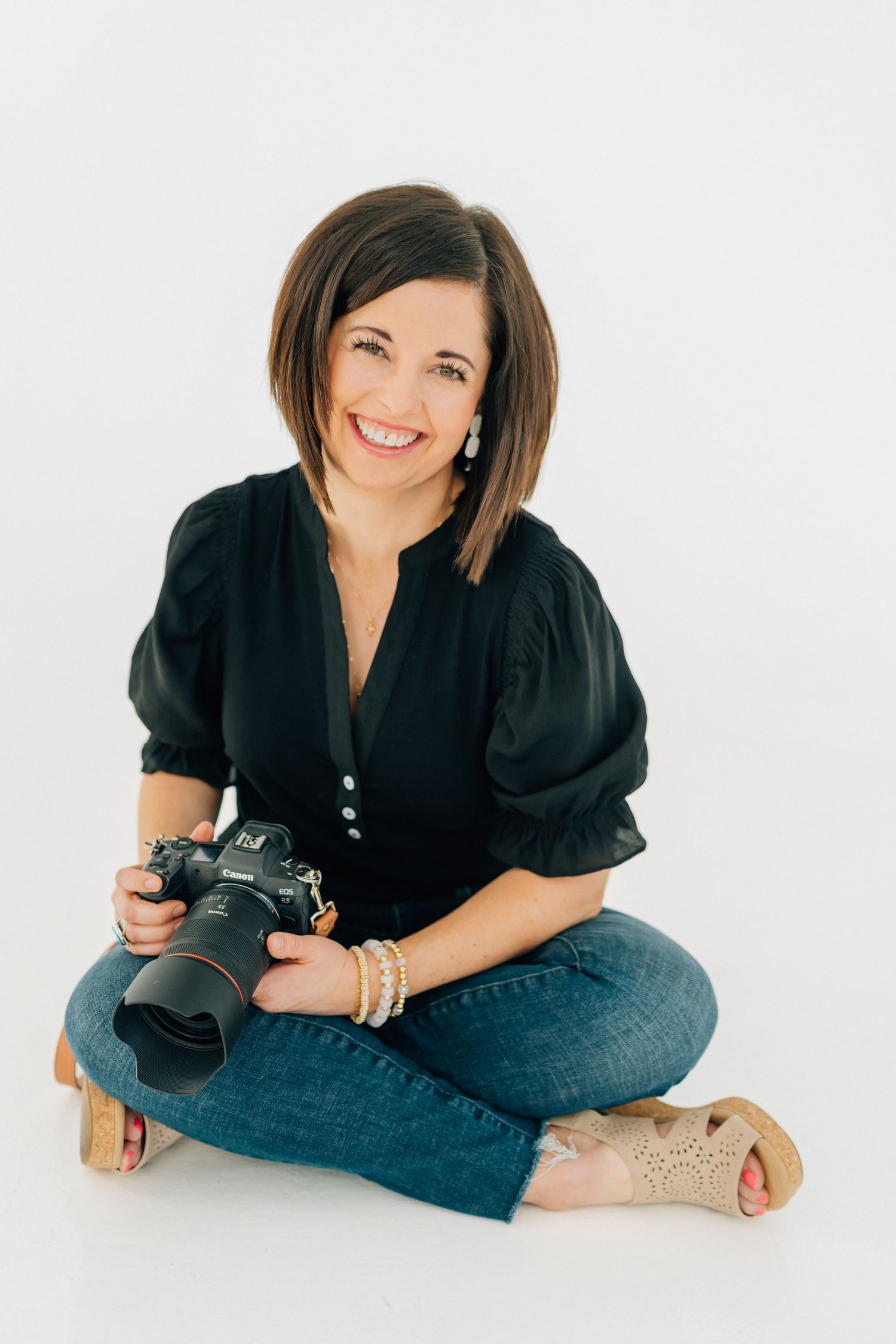 Professional photographer in black top and jeans poses casually with camera on white background in studio setting.