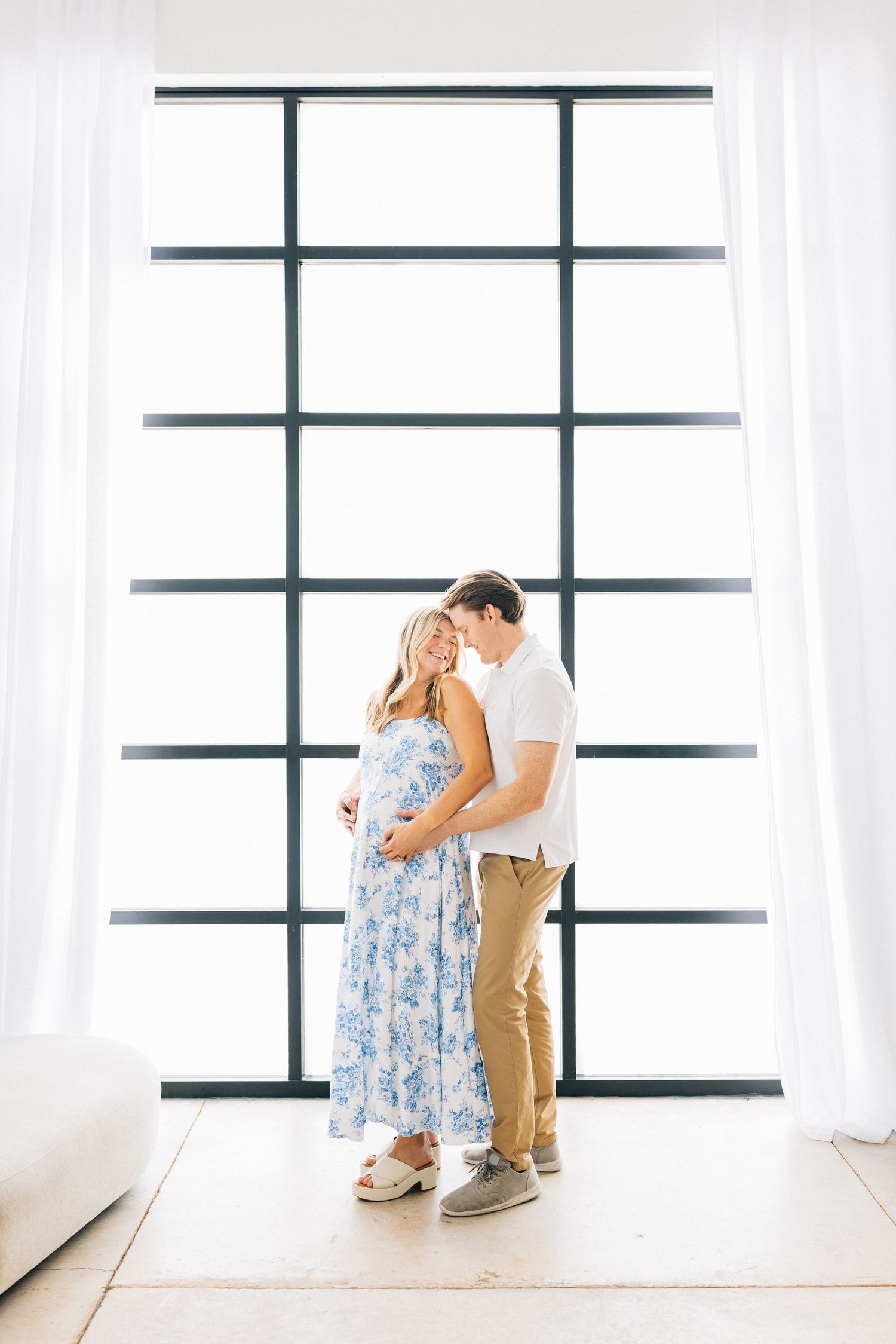 Someone wearing a blue floral dress and khaki pants embraces in a sunlit room with large window panels.