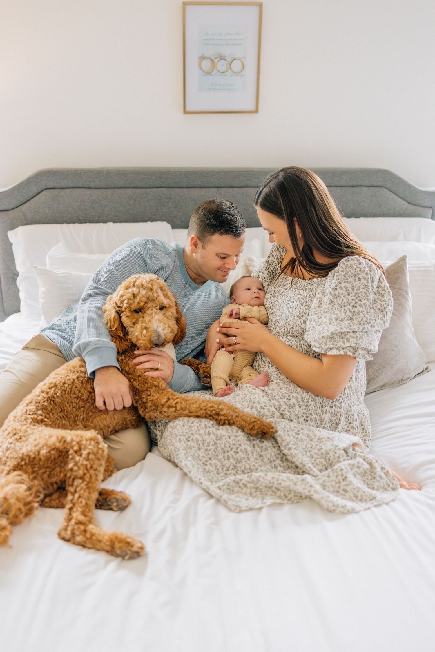 Family relaxing together on white bedding with their goldendoodle dog in a cozy bedroom setting.