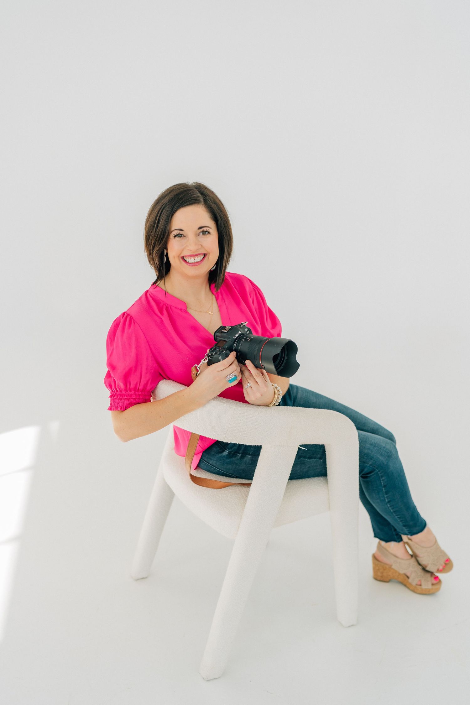 A person in a bright pink top and jeans sits casually on a white chair against a white background.
