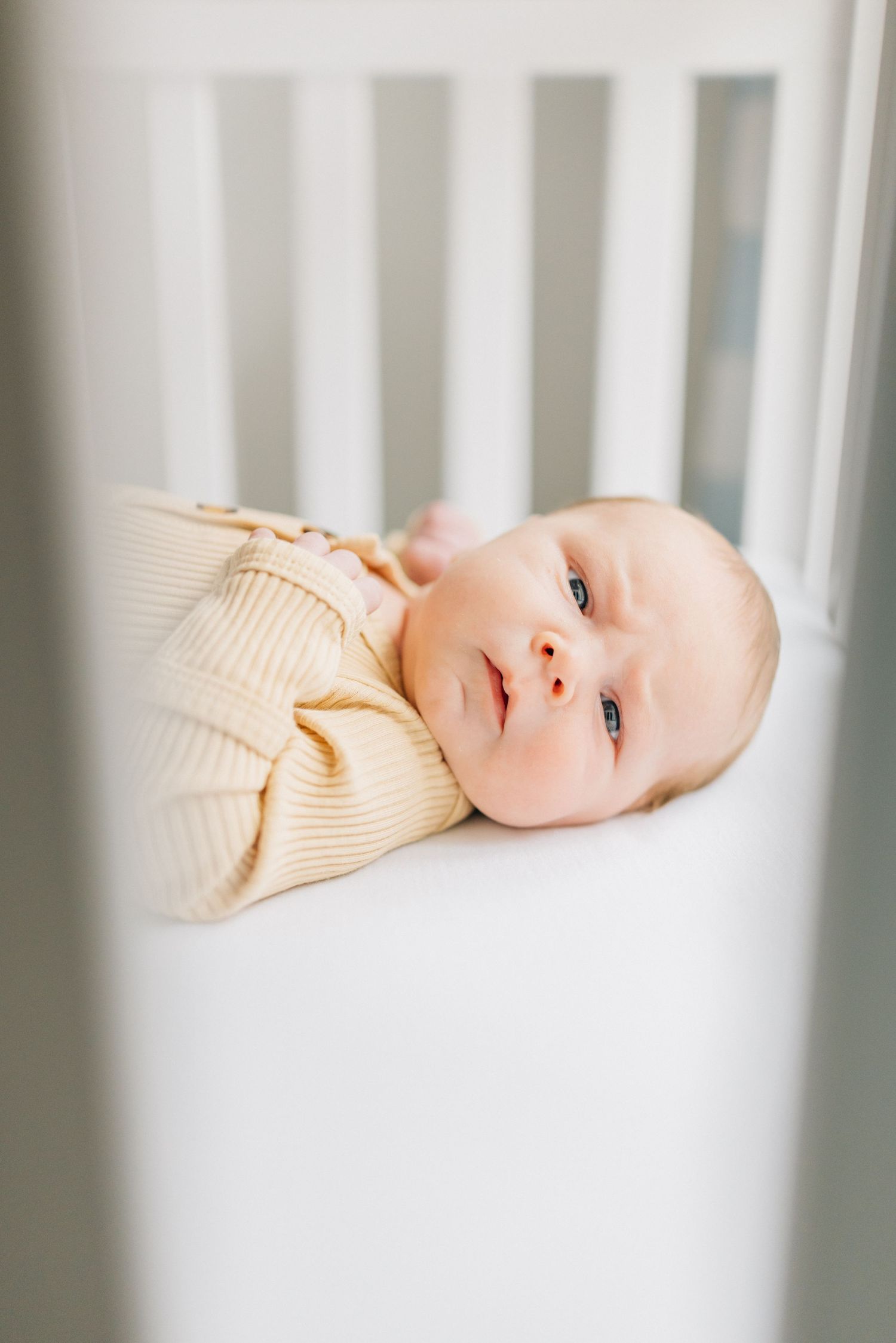 Baby in cream sweater lying in white crib looking up.