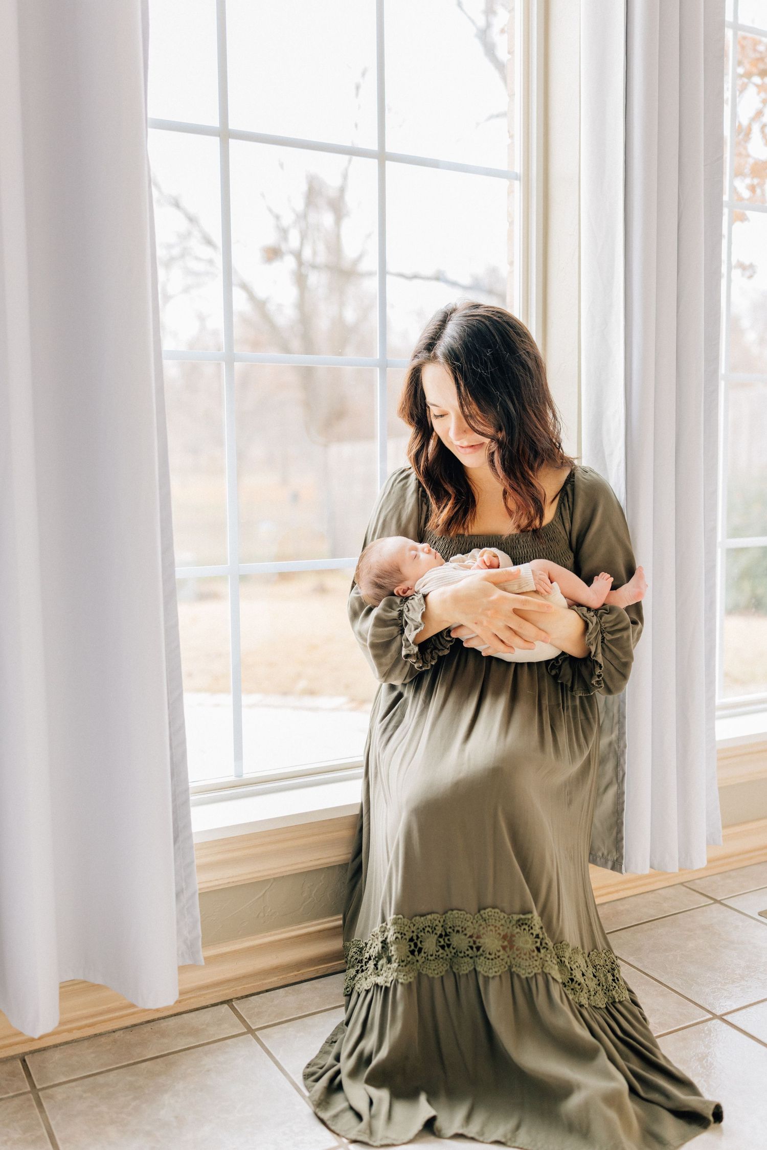 A person in an olive green maxi dress kneels by a bright window while holding a small white bundle.