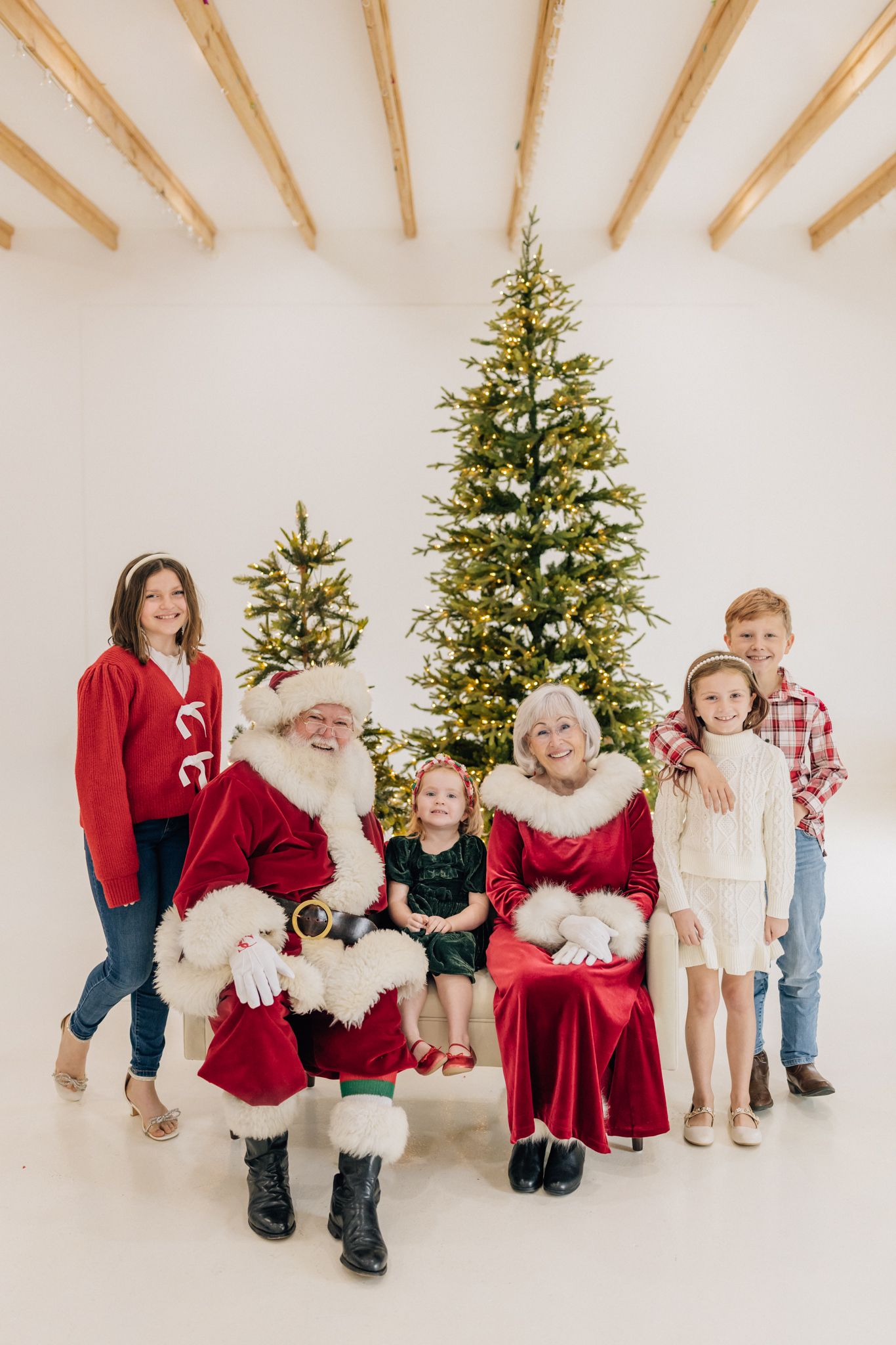 Family posing with Santa and Mrs. Claus in front of a lit Christmas tree in a bright white studio.
