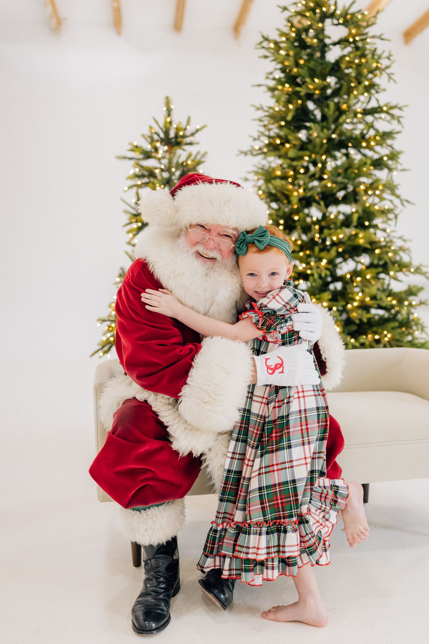 A young girl in plaid pajamas hugs Santa Claus beside a lit Christmas tree in a bright white studio setting.