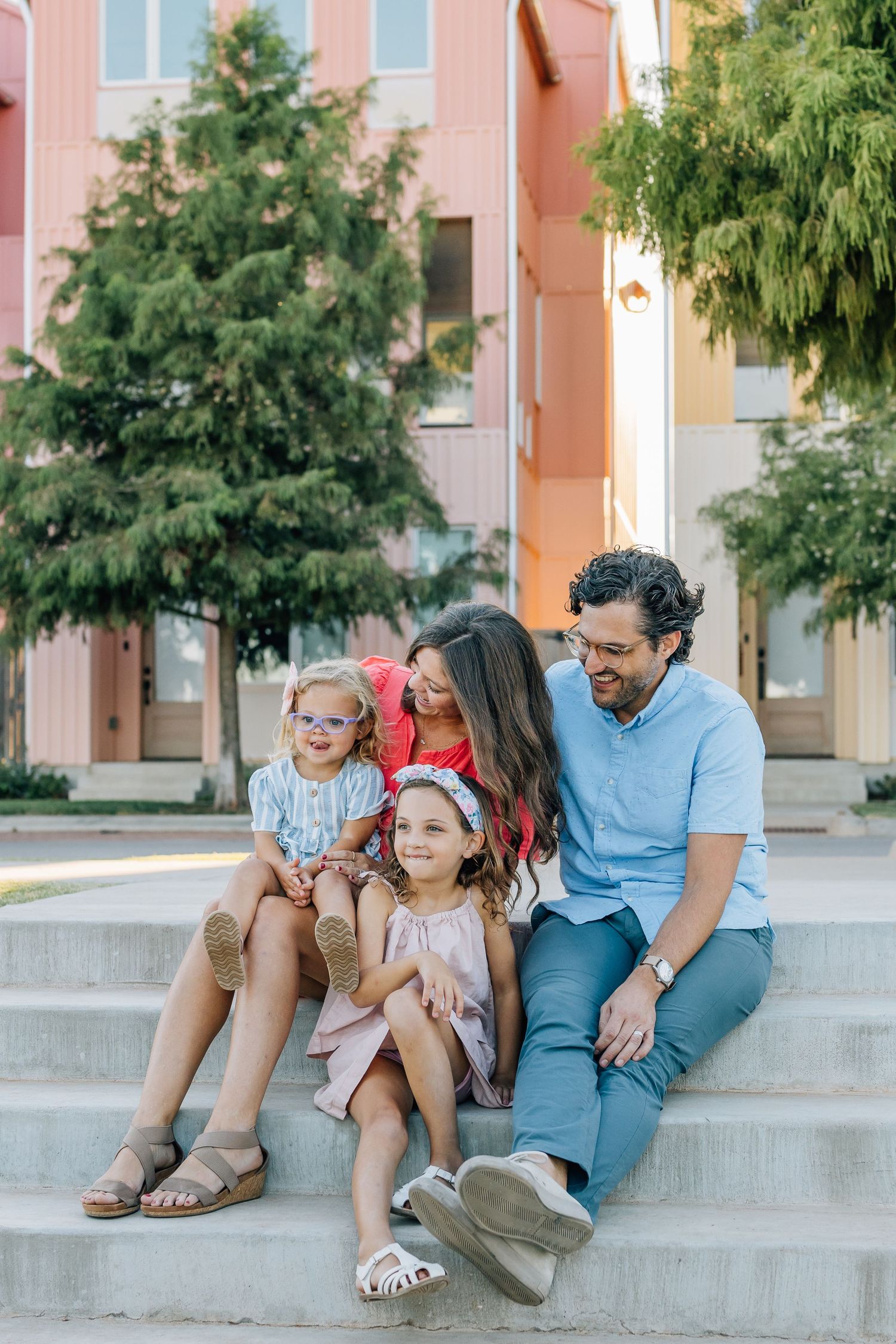 Family sitting together on concrete steps outside pink building on sunny day.