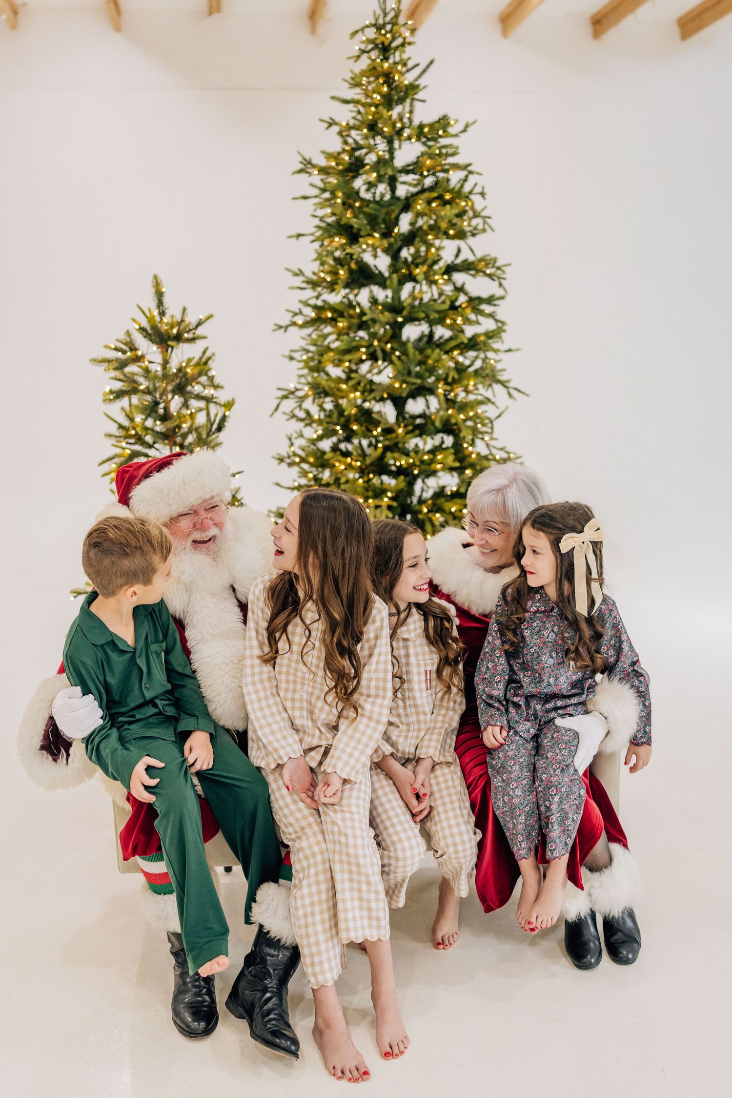 Santa Claus sits with four children in pajamas in front of a lit Christmas tree in a bright white studio setting.
