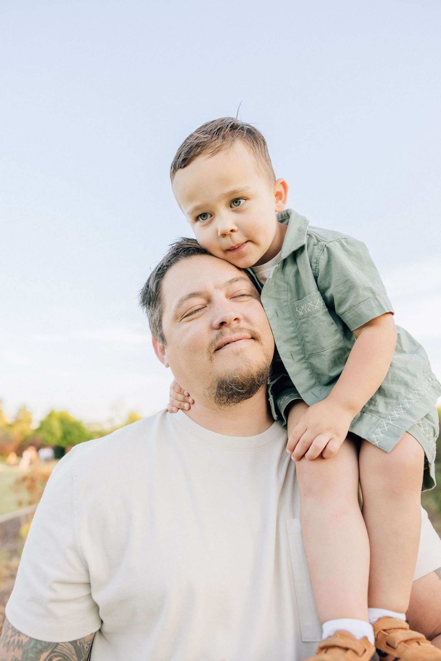 A tender outdoor moment captured between a child riding on shoulders and an adult in a white t-shirt on a bright sunny day.