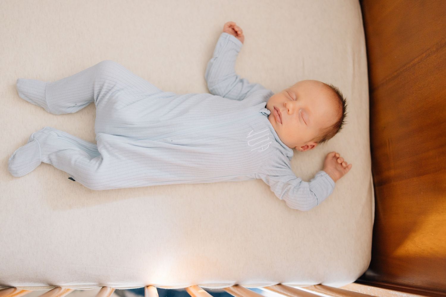 Newborn baby in white sleepwear rests peacefully on a white pillow against a wooden headboard.