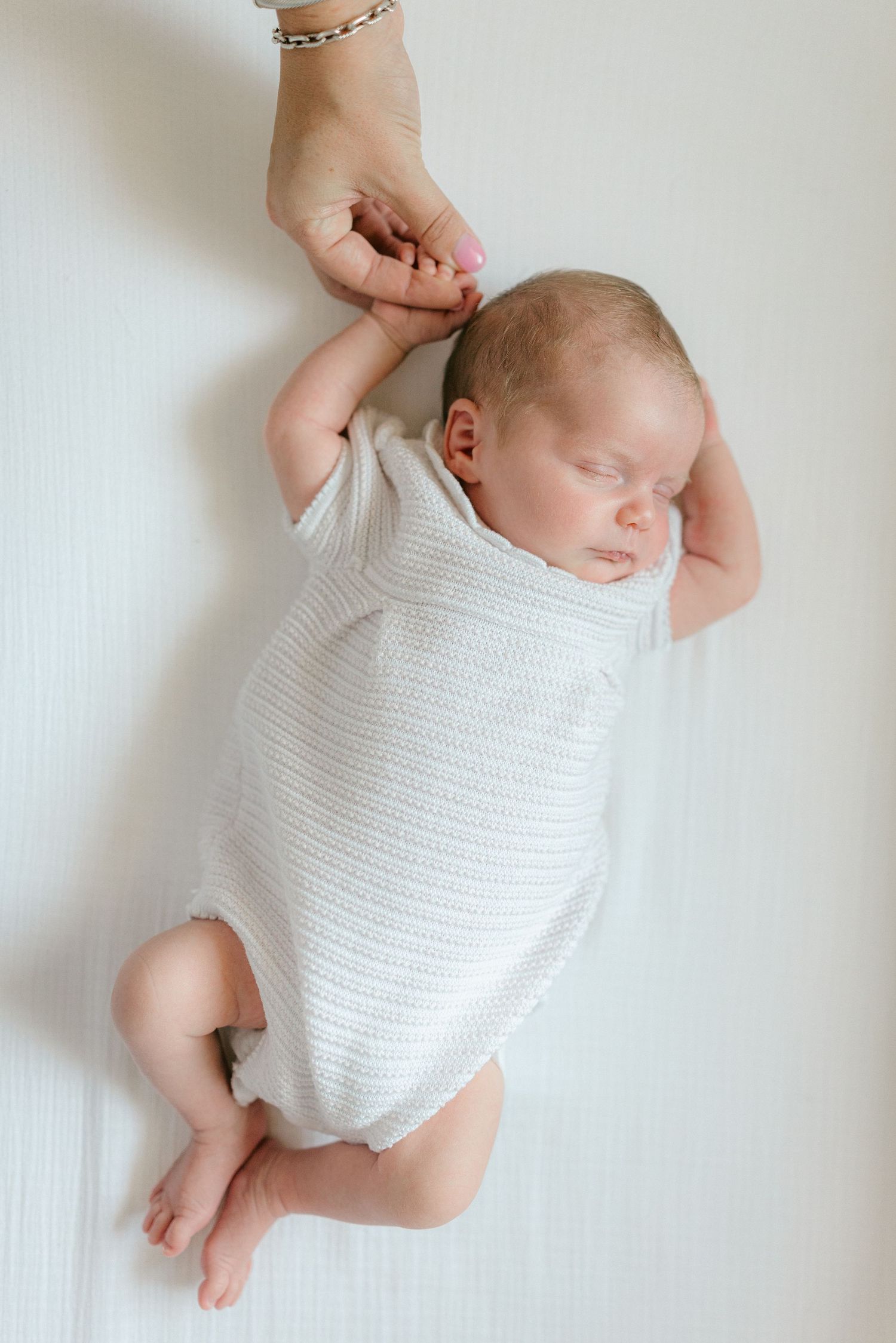 Sleeping newborn baby in white onesie gently holding an adult's finger on a white background.