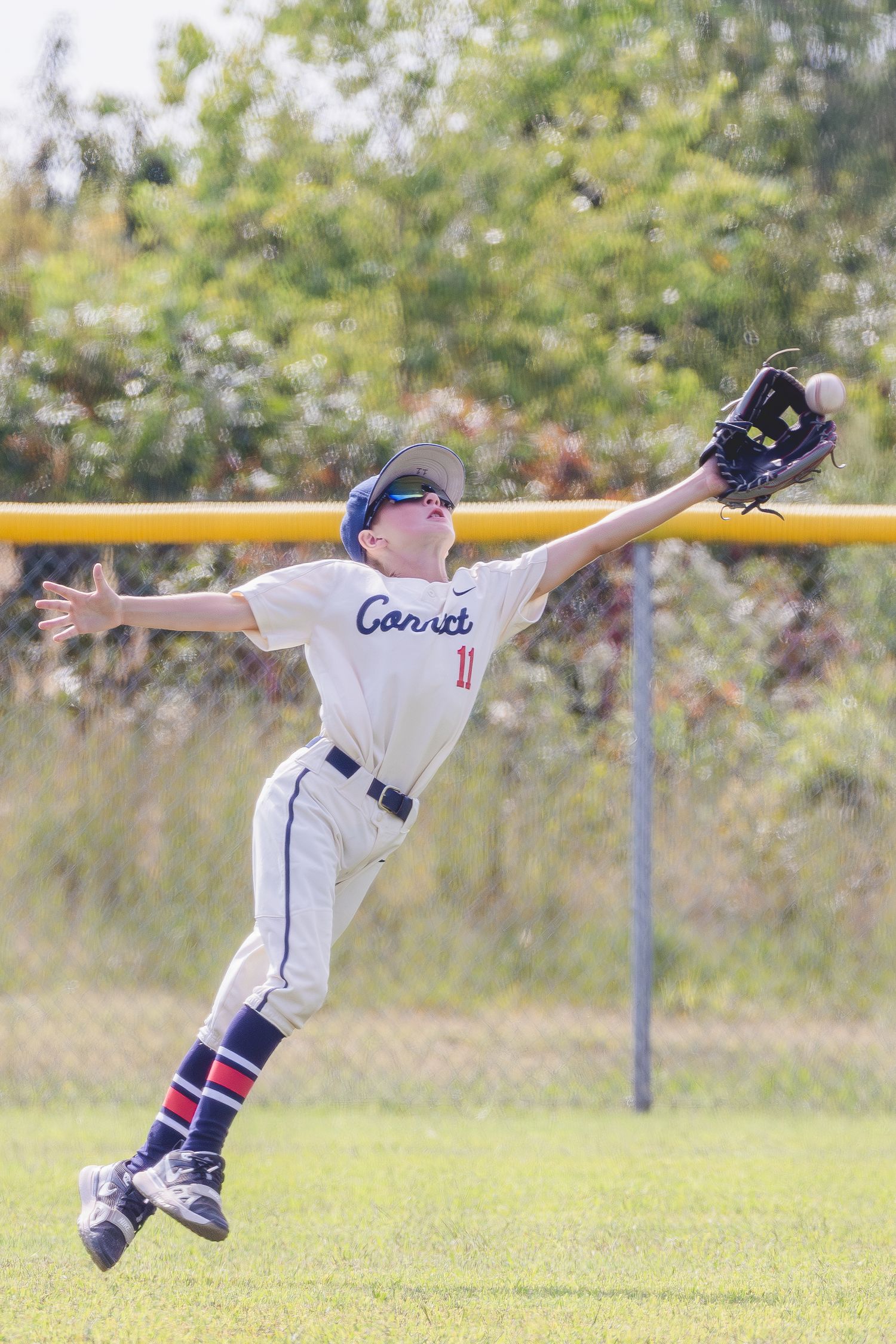 Youth baseball player leaping to catch a fly ball during a game on a sunny day at a grass field.