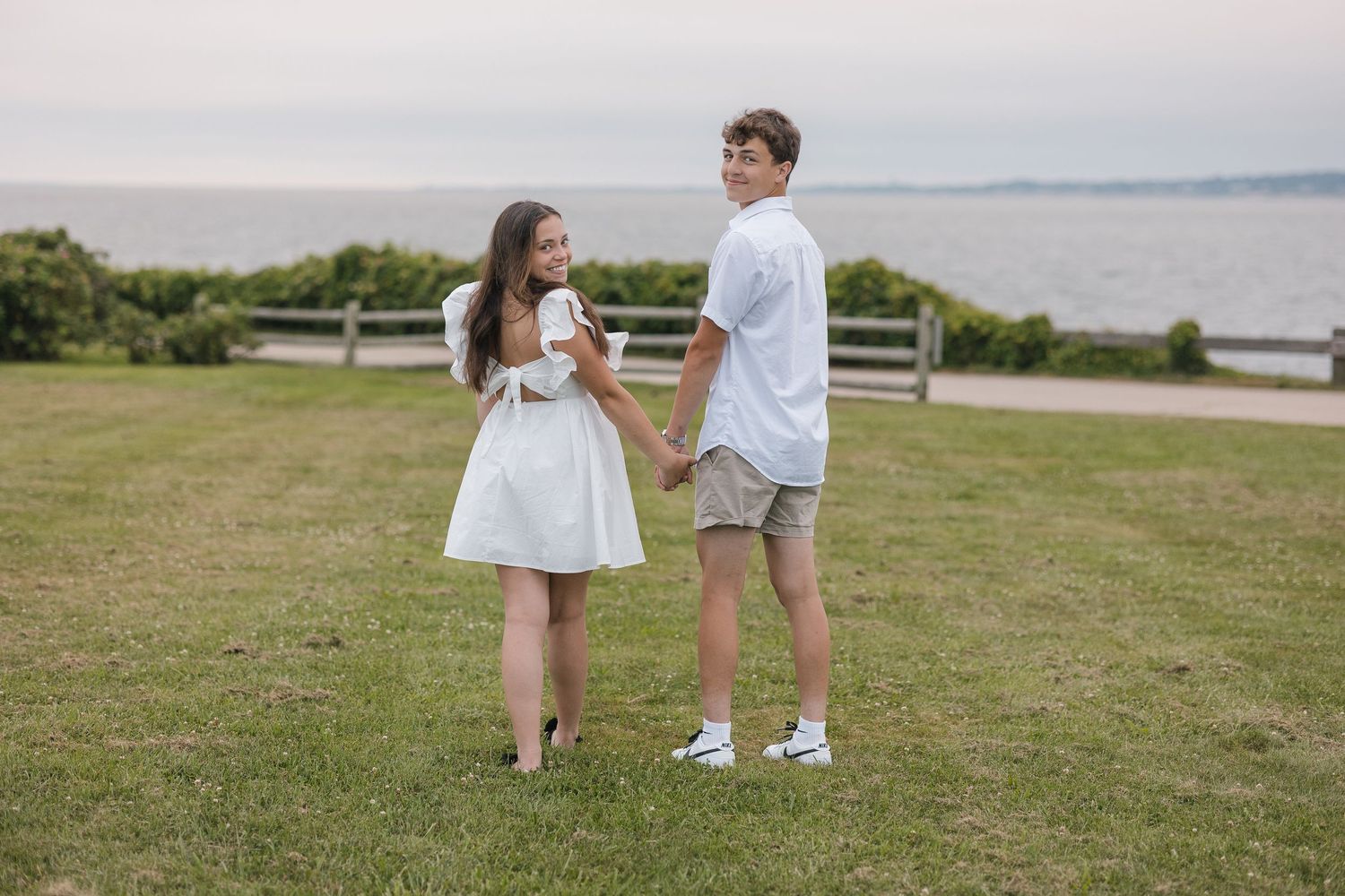 A couple in white summer outfits holds hands while standing on a grassy hill overlooking the ocean.