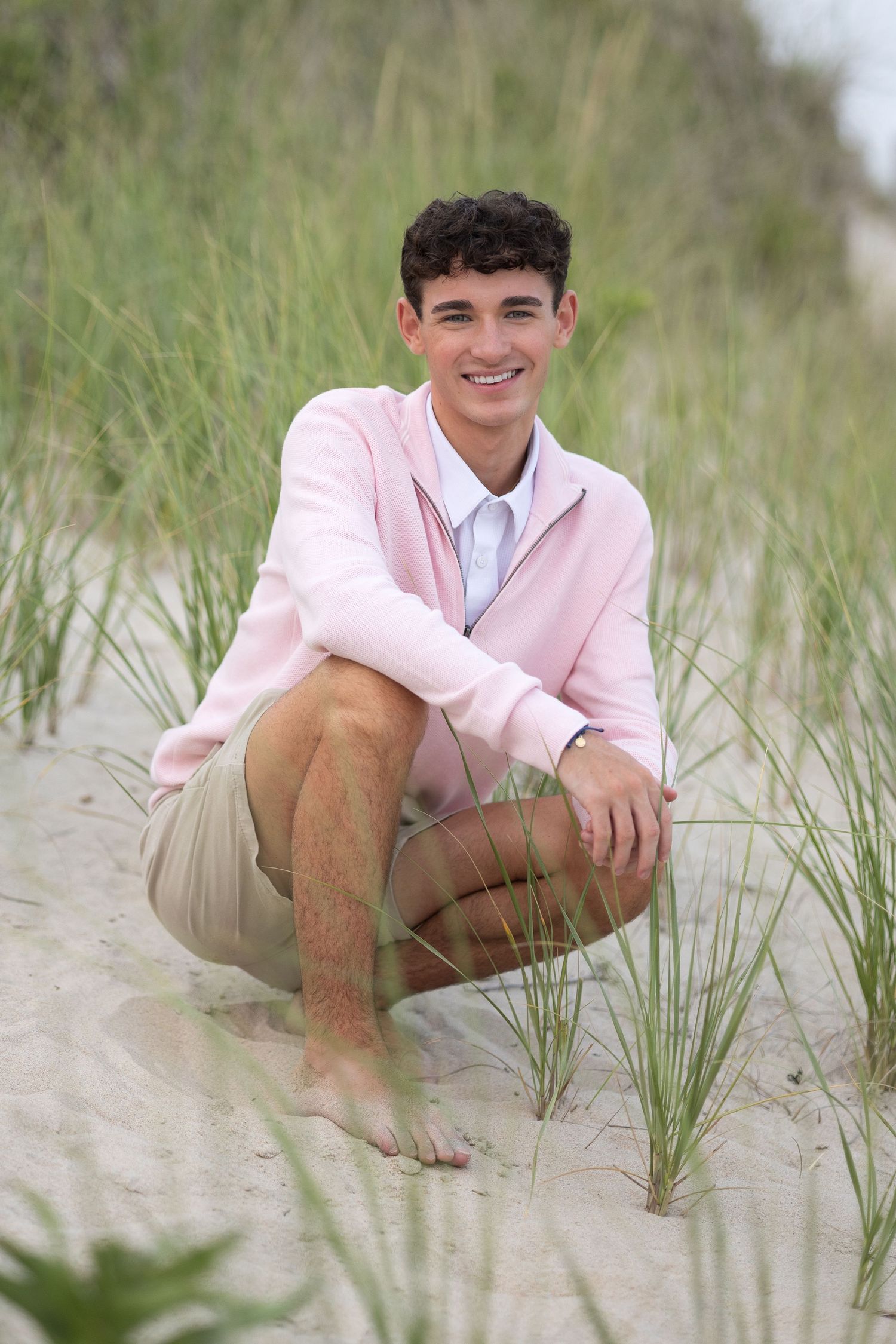 A young person in a pink blazer and khaki pants poses casually on a sandy beach with sea grass.