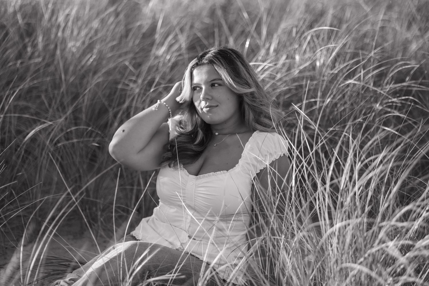 Black and white portrait of woman in white dress sitting among tall grass in a dreamy outdoor setting.