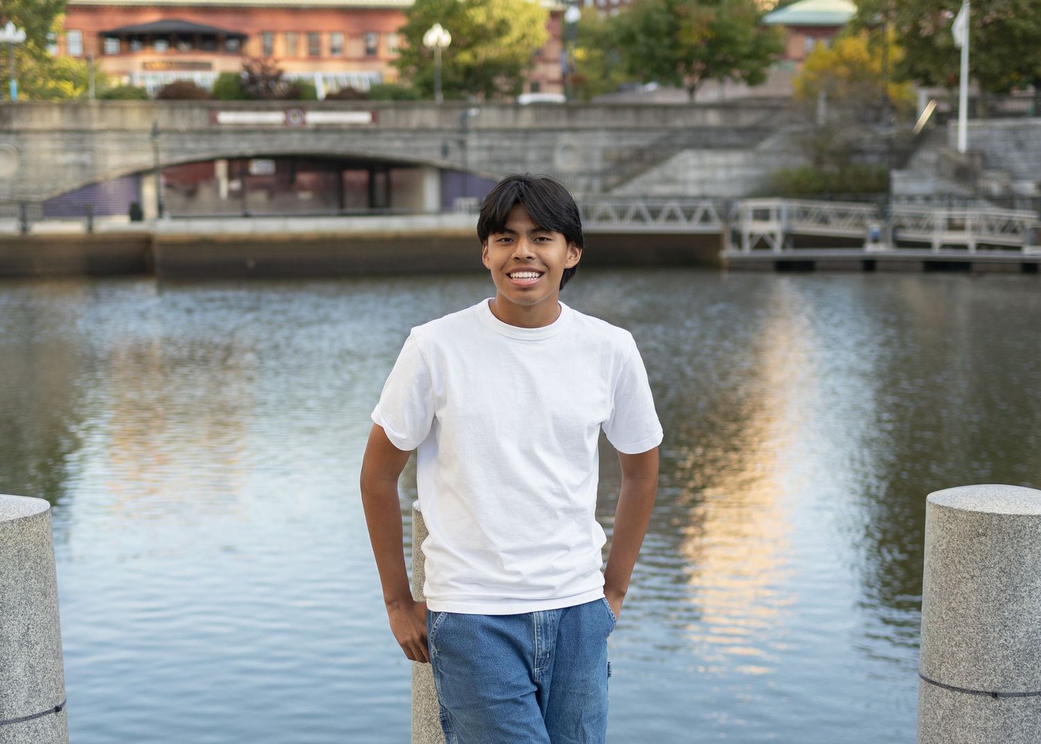 Person in white t-shirt stands by waterfront with urban architecture in background.