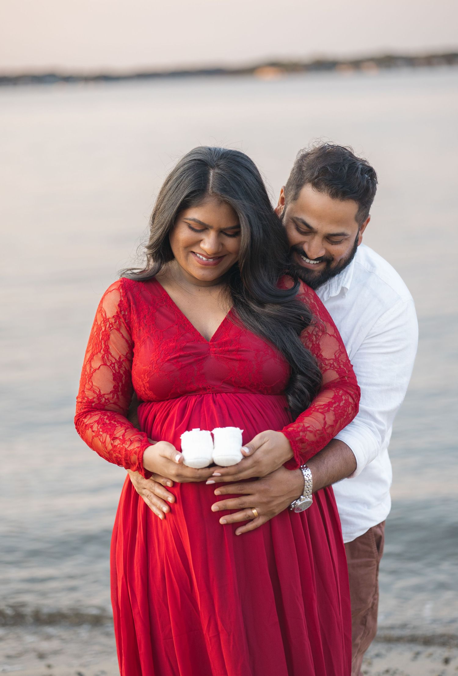 Couple holding tiny white baby shoes against beachfront backdrop.