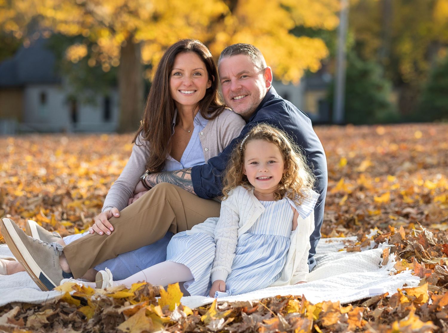 Family lying in colorful autumn leaves during outdoor photo session with golden fall foliage in background.