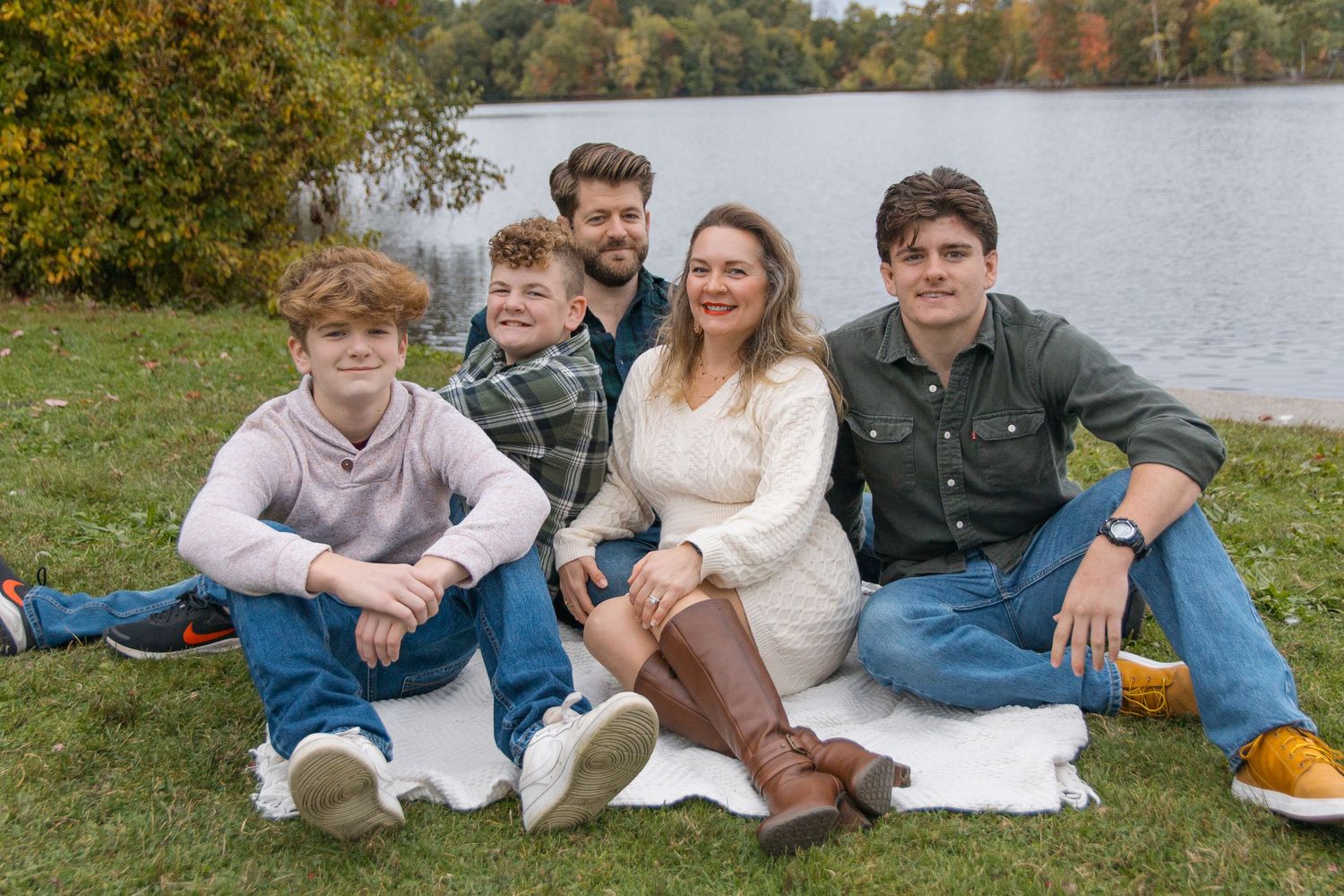 A group relaxes on a white blanket by a lake during autumn.