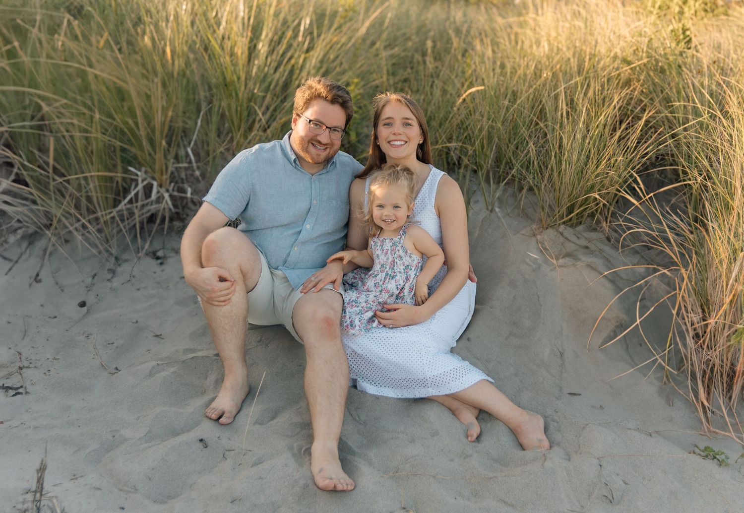 A couple sits with their young child on a sandy beach surrounded by tall dunes at sunset.