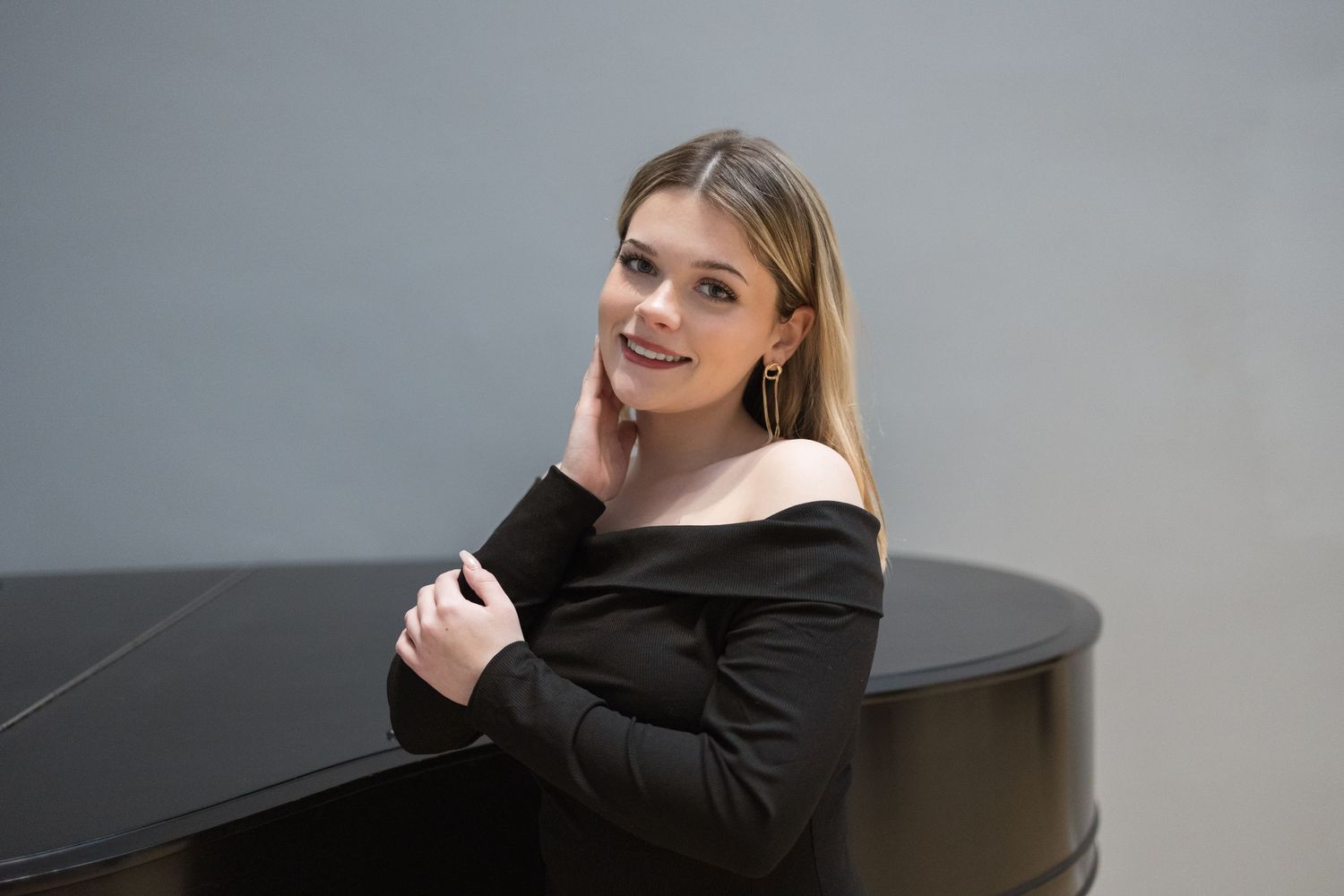 Woman in black off-shoulder dress poses beside black piano in gallery space.