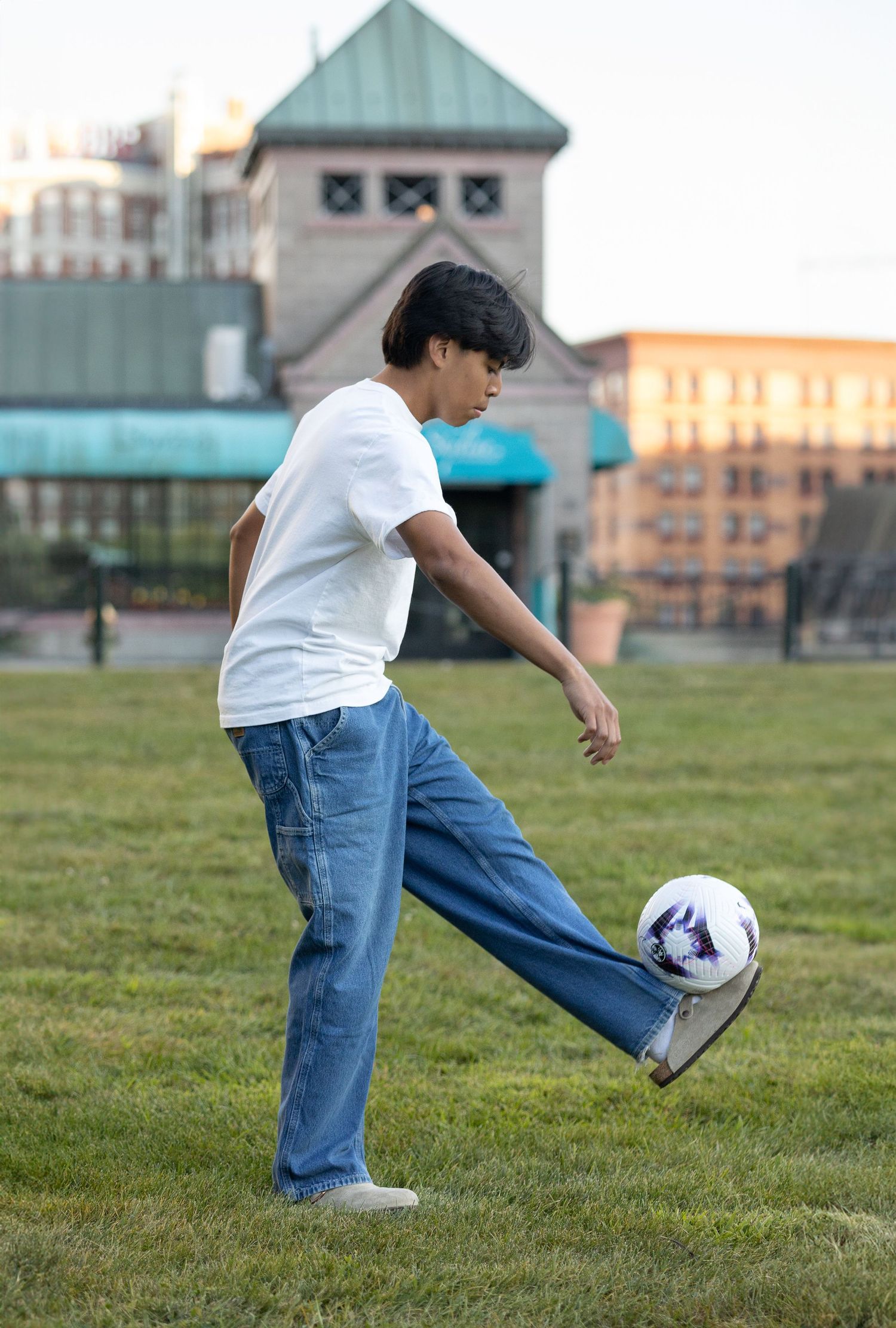 Person practicing soccer moves with ball on grass field with urban building in background.
