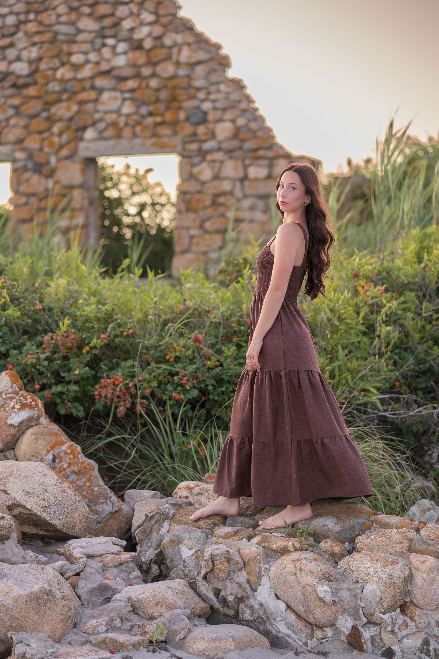 A woman in a brown dress poses on rocky terrain near a stone wall at sunset.