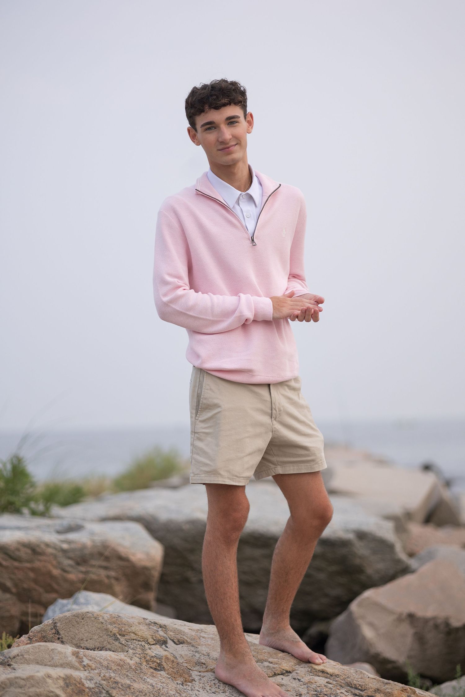 Model poses on rocky beach wearing light pink sweater and khaki shorts in coastal photoshoot.