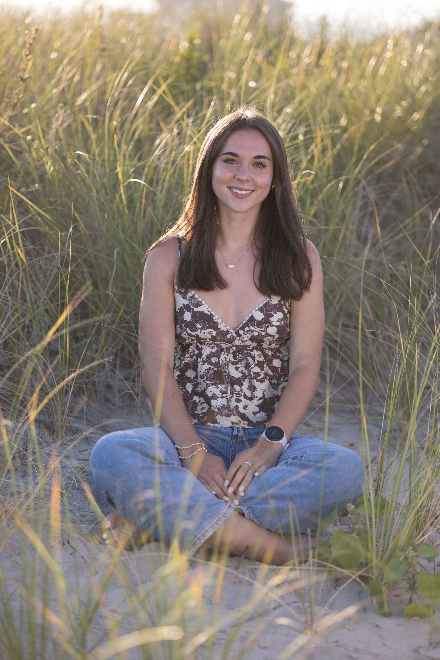 A person in a floral tank top and jeans sits cross-legged among tall beach grass at sunset.