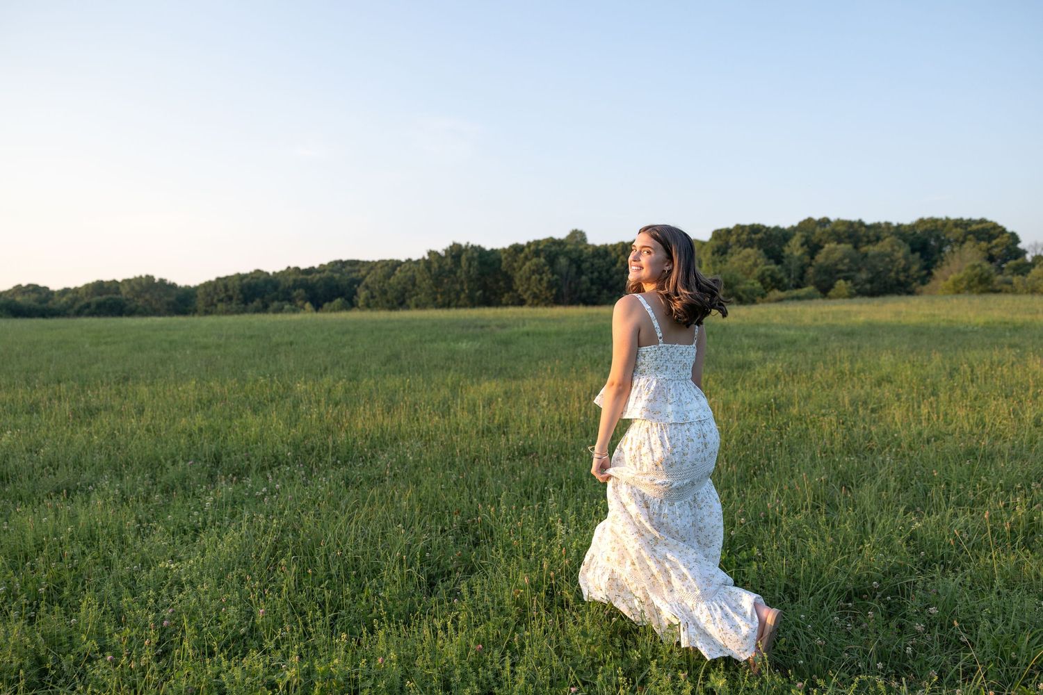 HS senior in a flowing white dress runs through a lush green field at Chase Farm Rhode Island golden hour with trees in the background.