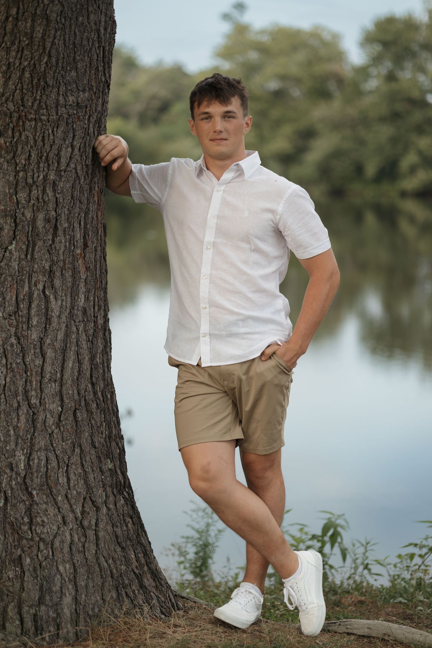 Someone poses by a large tree trunk near a lake wearing a white shirt and khaki shorts.