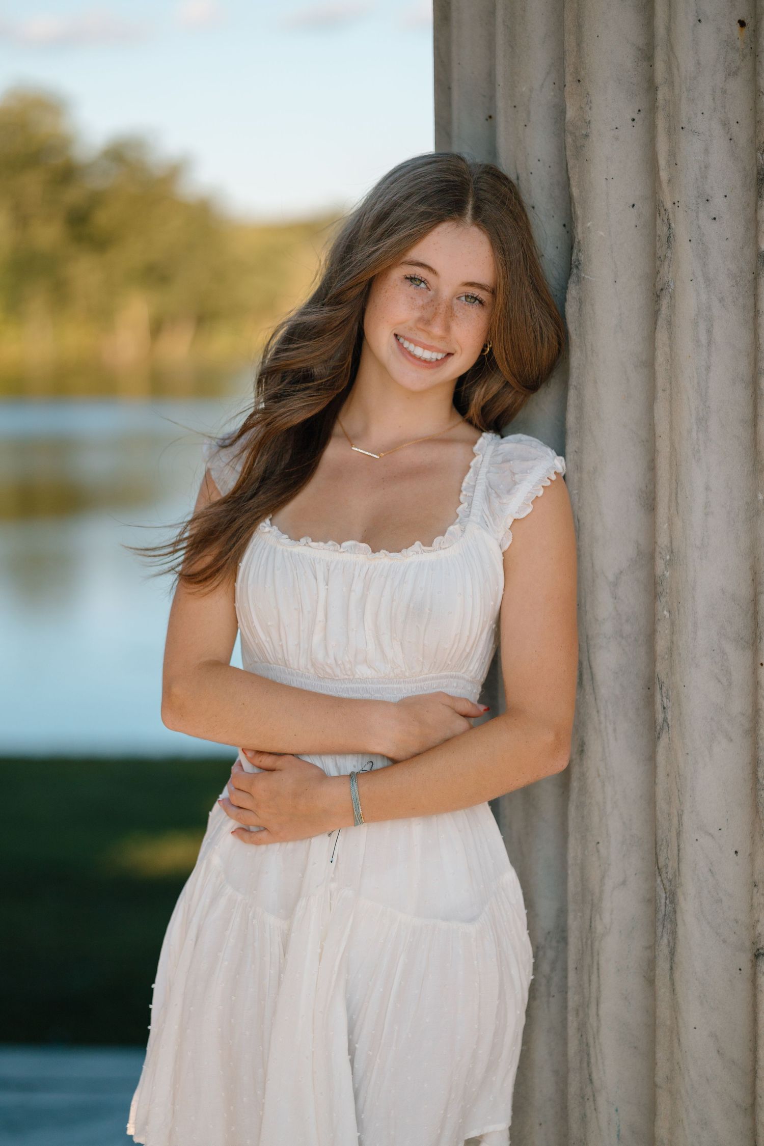 Young woman in white sundress poses against column by lake during golden hour outdoor portrait session.
