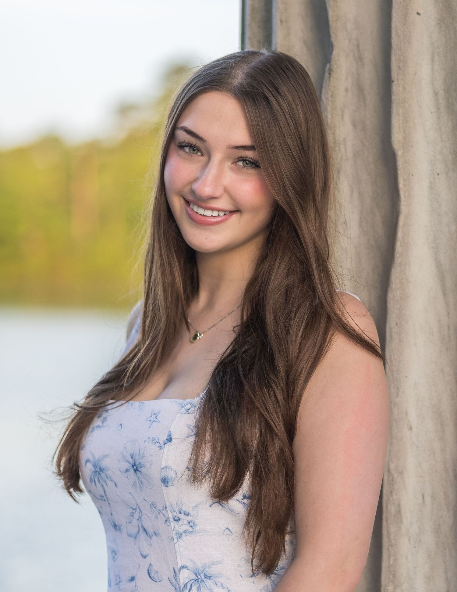 A person in a light blue floral dress poses outdoors against a natural background with water and trees.