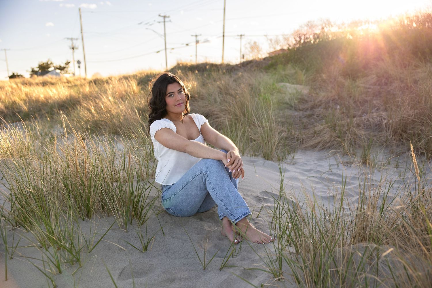 Woman in white top and jeans sitting on sandy dunes at golden hour with beach grass at beach in Narragansett, RI