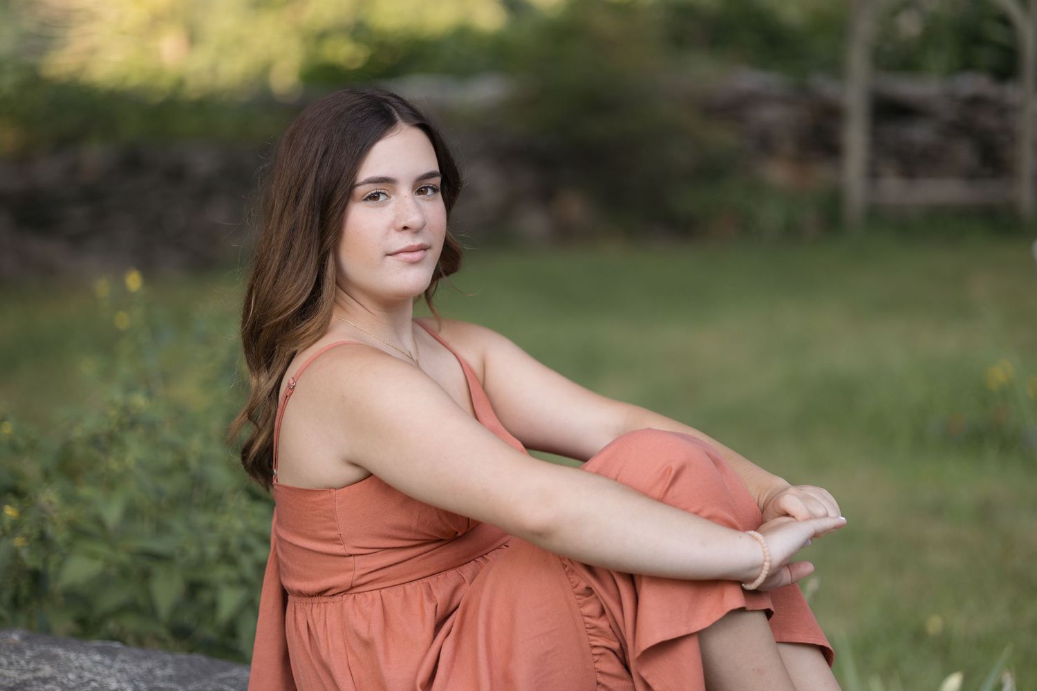 Woman in flowing coral dress sits outdoors on stone wall in garden setting during golden hour.