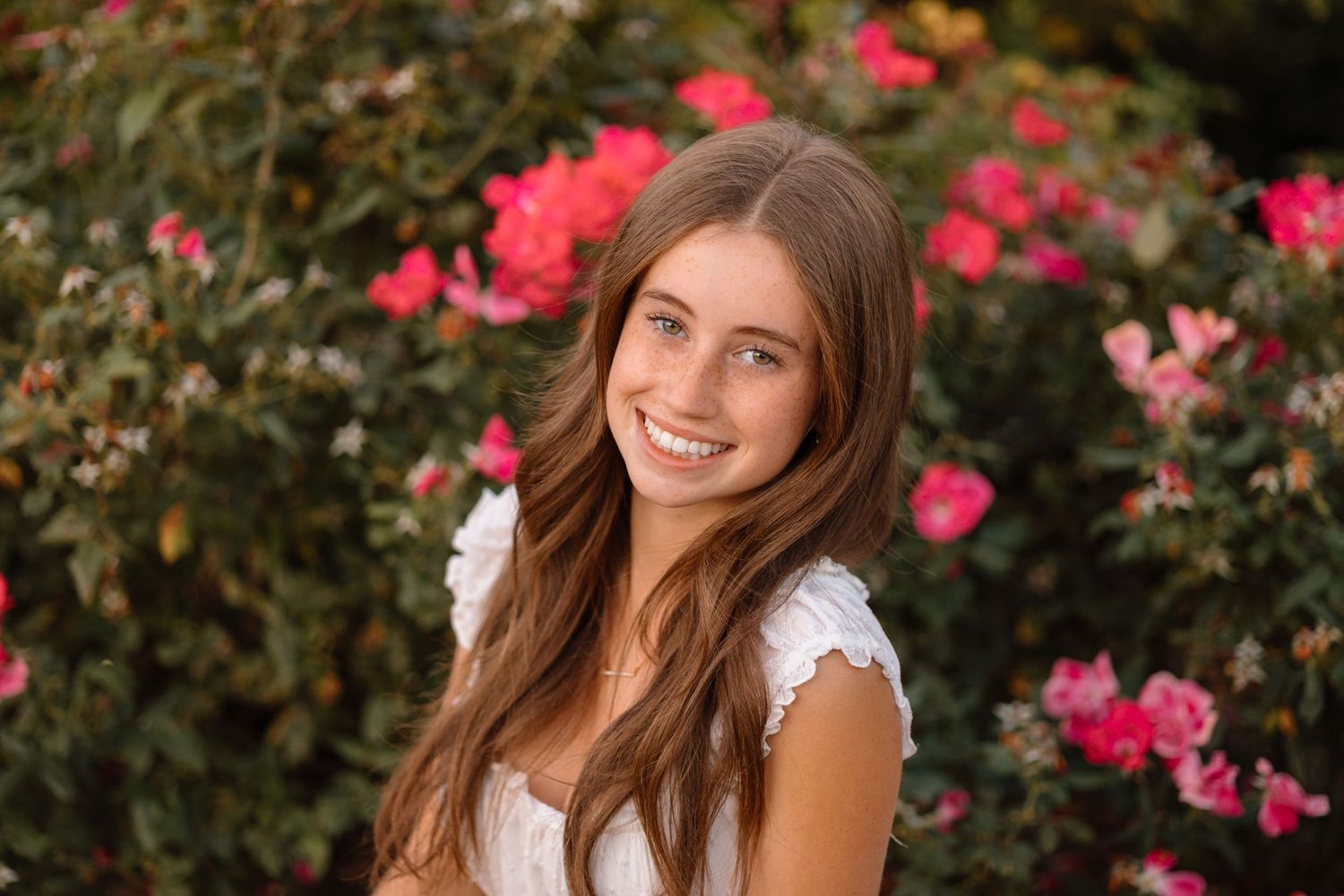 Woman in lace-trimmed white dress poses next to vibrant pink rose bushes during summer photo session.