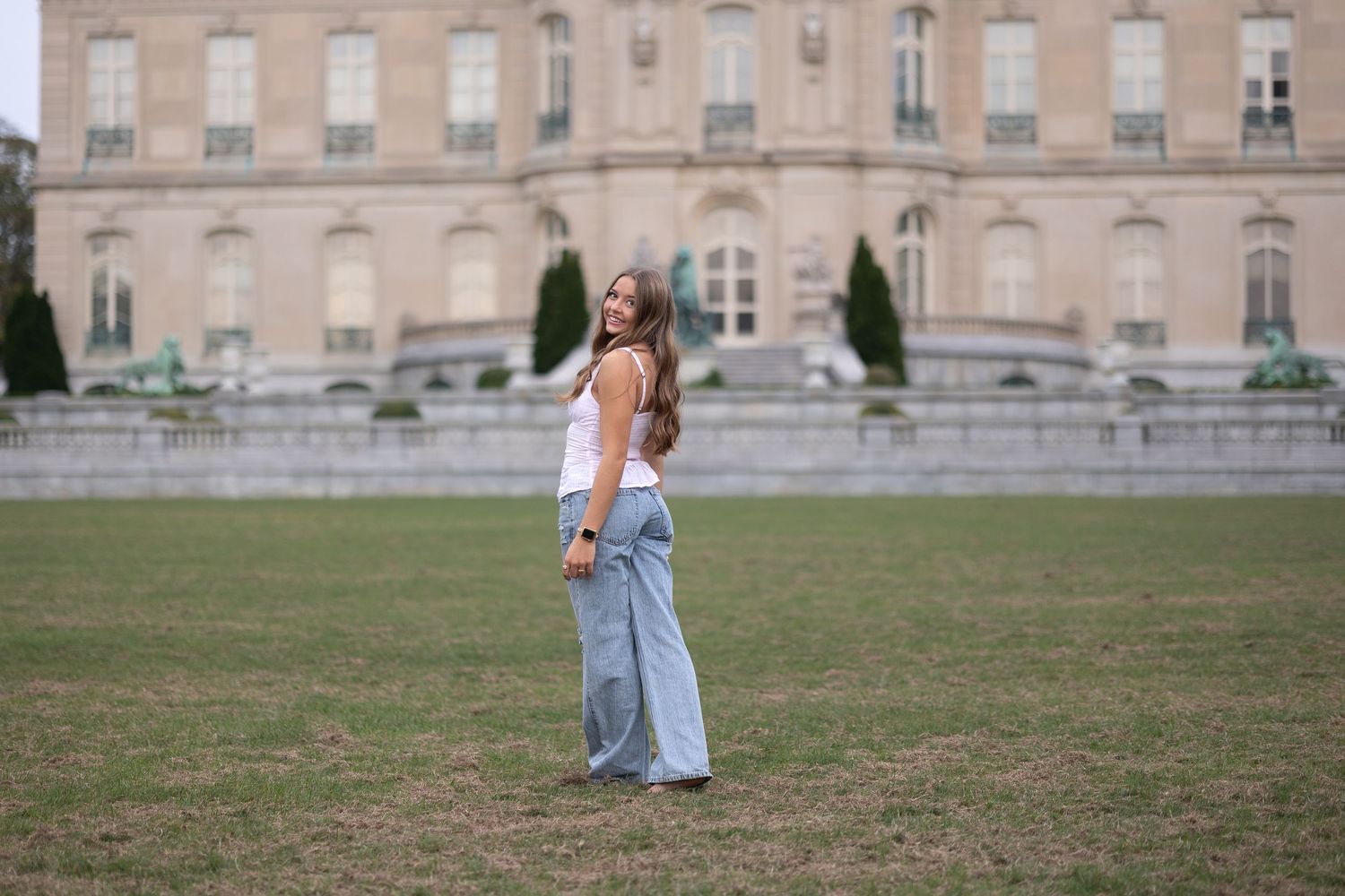A person in casual attire poses on a manicured lawn in front of a grand historic mansion.