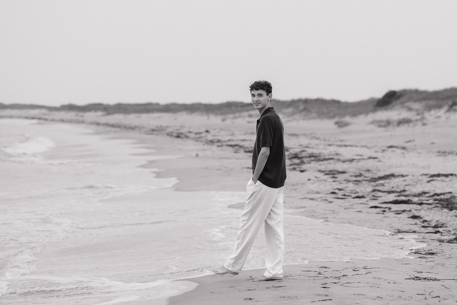 A person walks along a misty beach shoreline in white pants while waves roll onto the sandy coast.