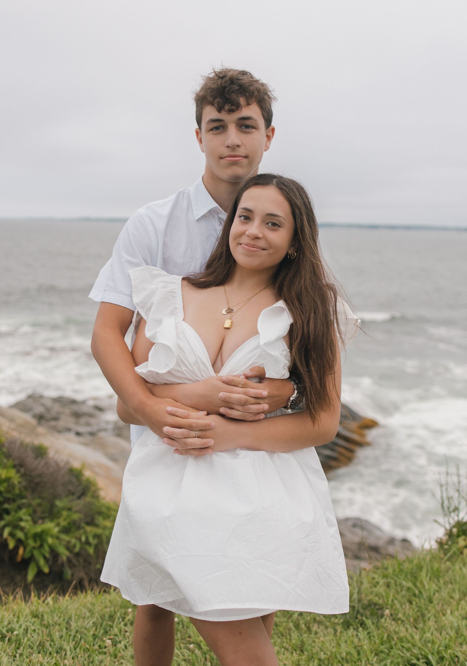 Couple in matching white outfits posing affectionately on grassy coastal overlook by the ocean.
