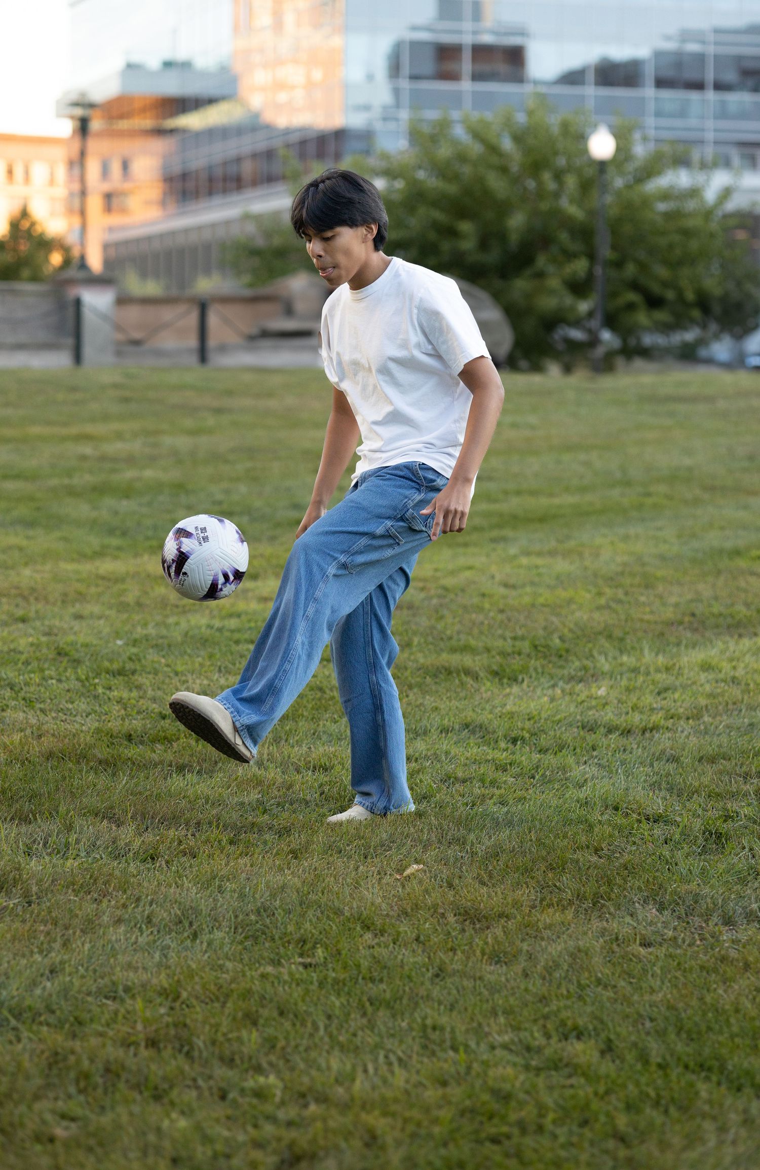 A person kicks a soccer ball on a grassy field during sunset with buildings in the background.