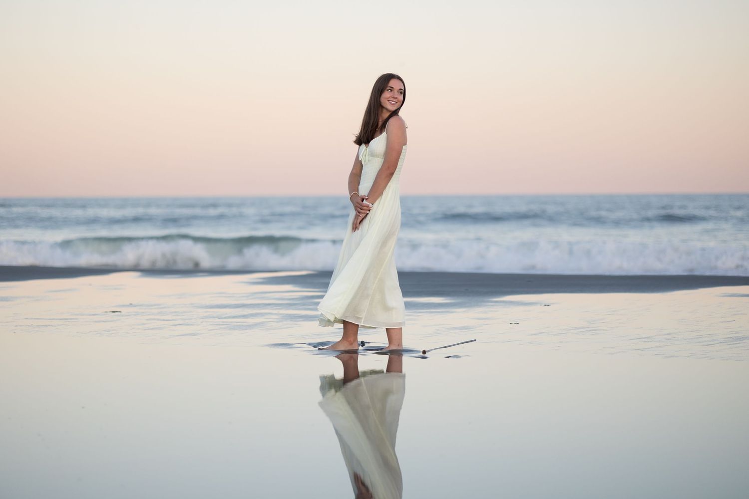 A person in a flowing white dress stands reflectively on a beach at sunset with waves in the background.