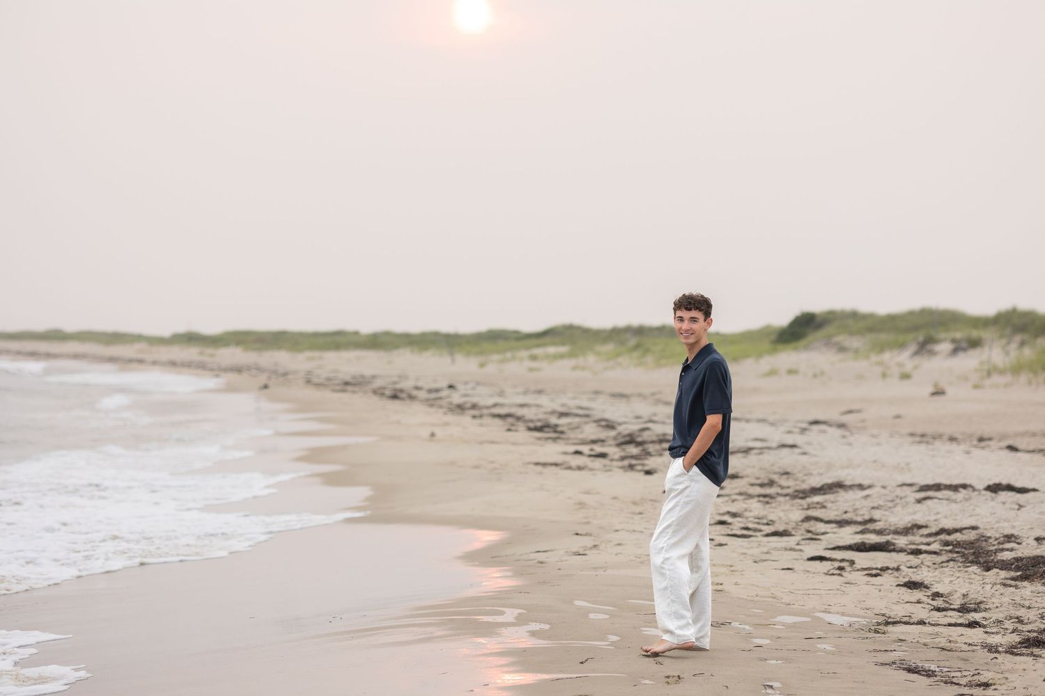 A person stands on a serene beach at sunset wearing a navy shirt and white pants along the shoreline.
