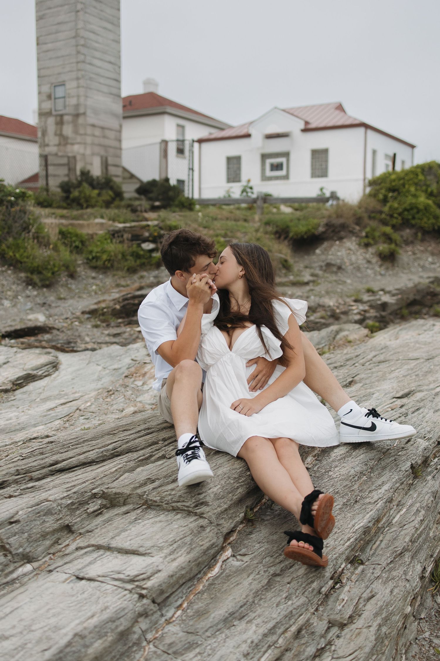 Couple in white outfits sharing intimate moments on rocky coastal terrain with houses in background.