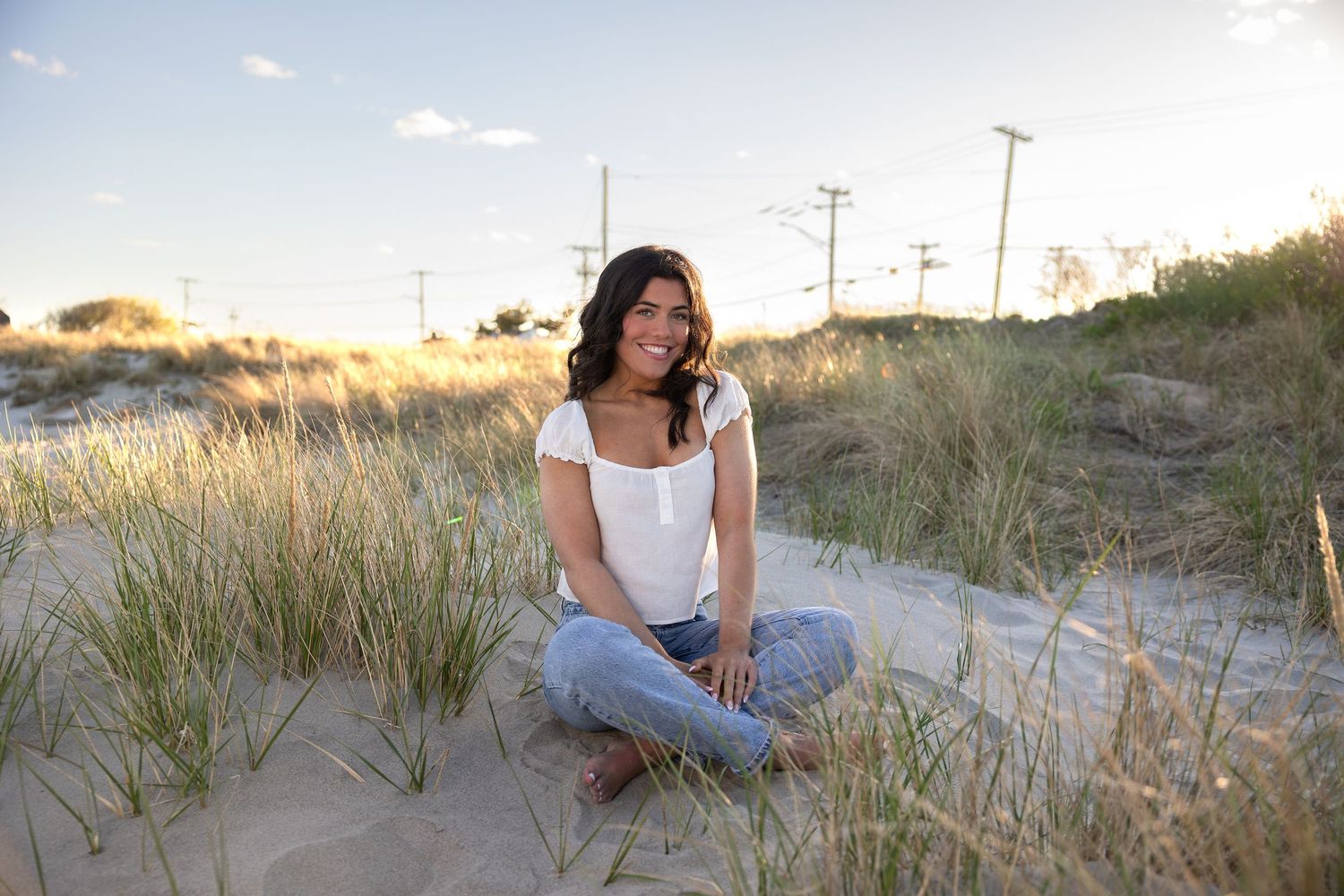Young woman sitting cross-legged on sandy beach dunes, smiling, wearing white top and jeans at golden hour.