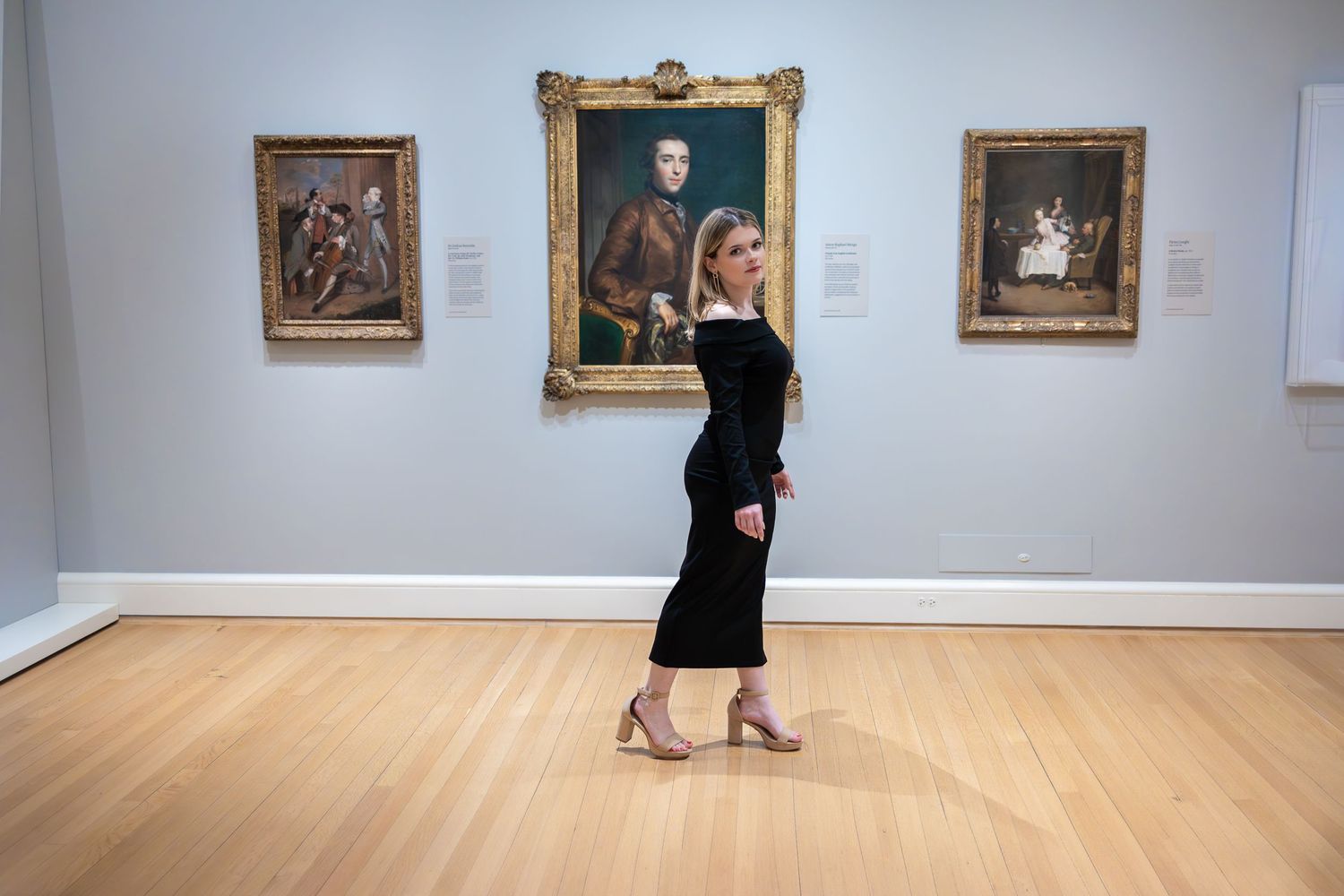 Person in long black dress walks through gallery displaying classical portraits.