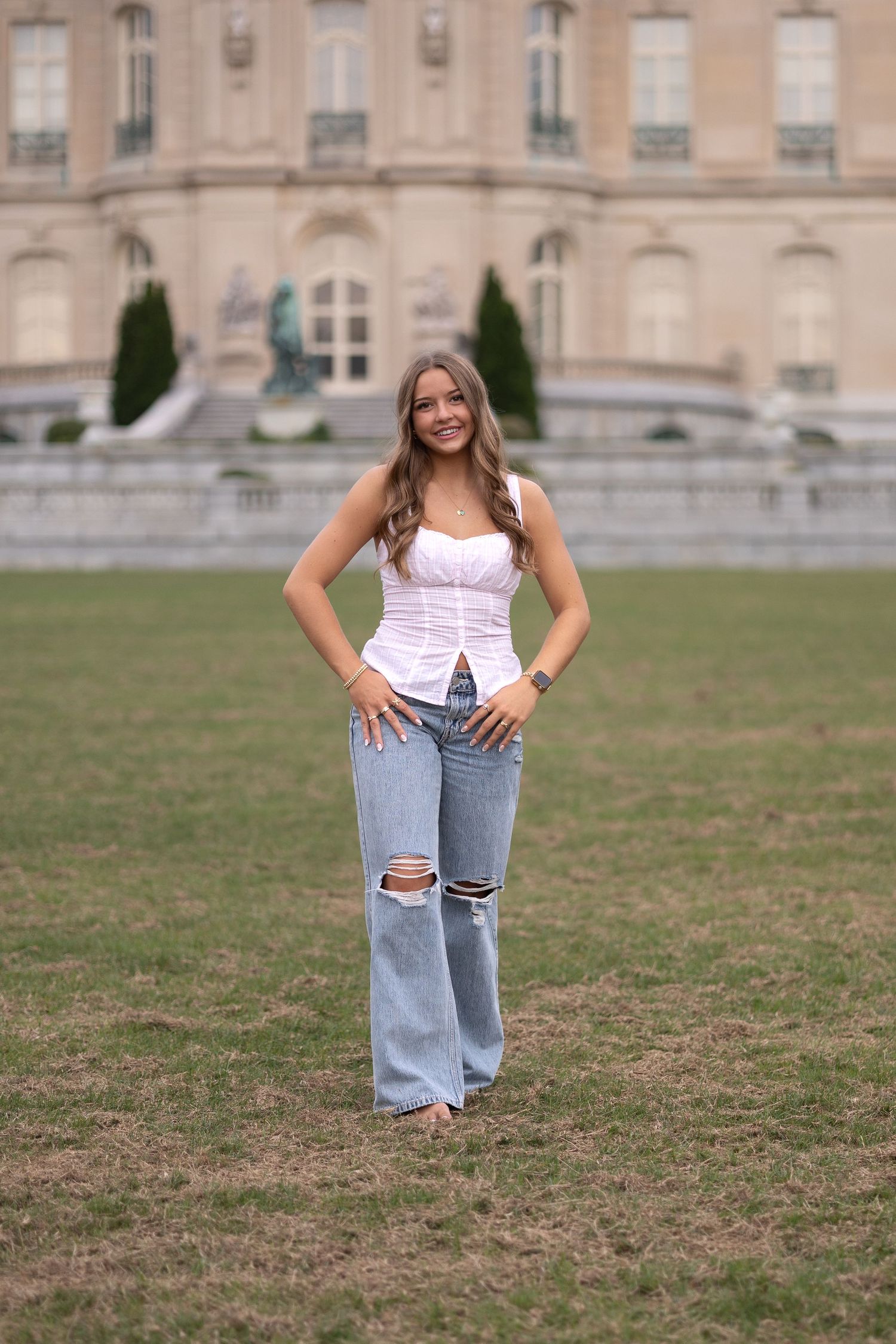 A person in ripped flare jeans and white corset top poses on grass in front of a grand historic building.