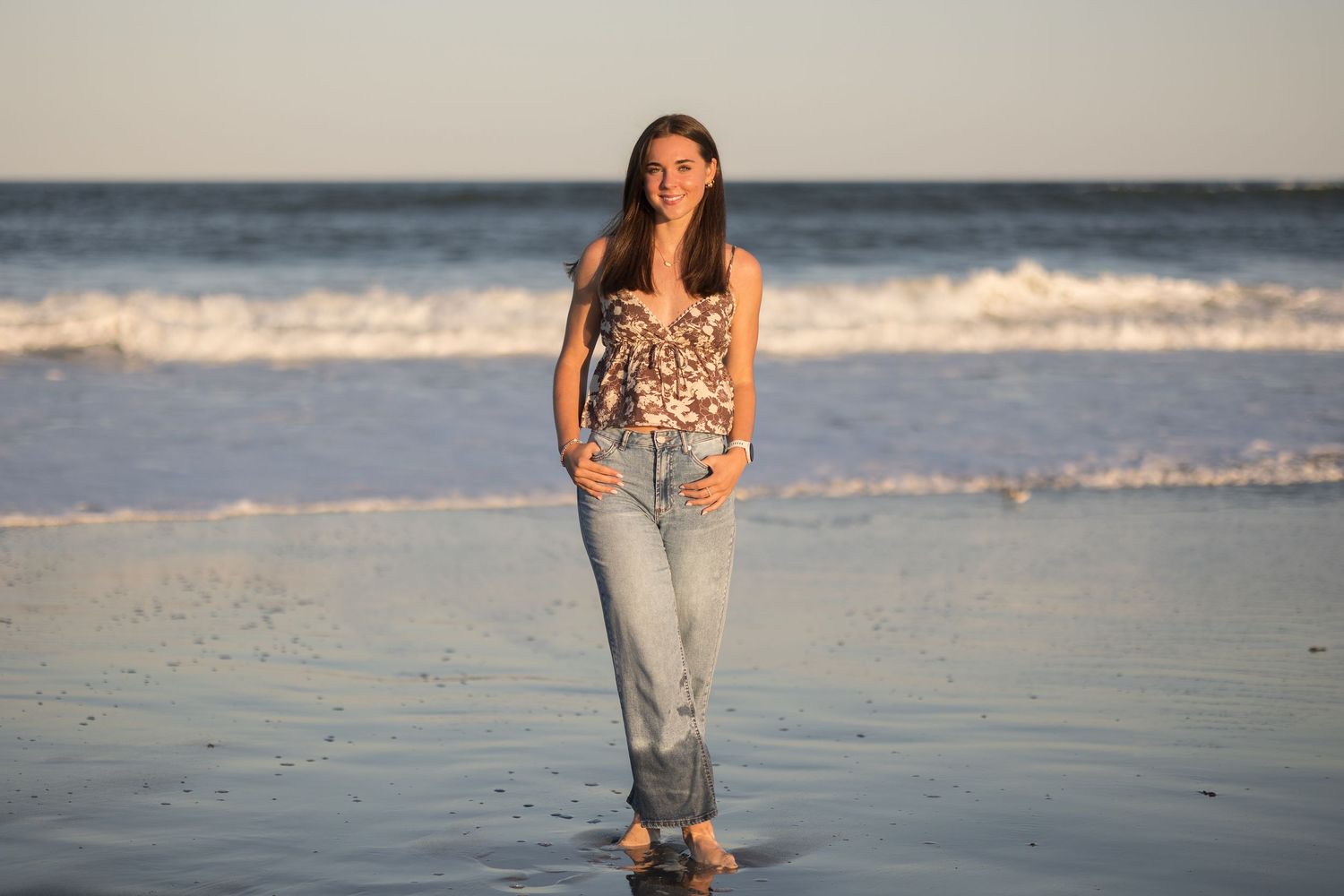 A person in casual attire walks along the shoreline at sunset while waves gently roll onto the sandy beach.