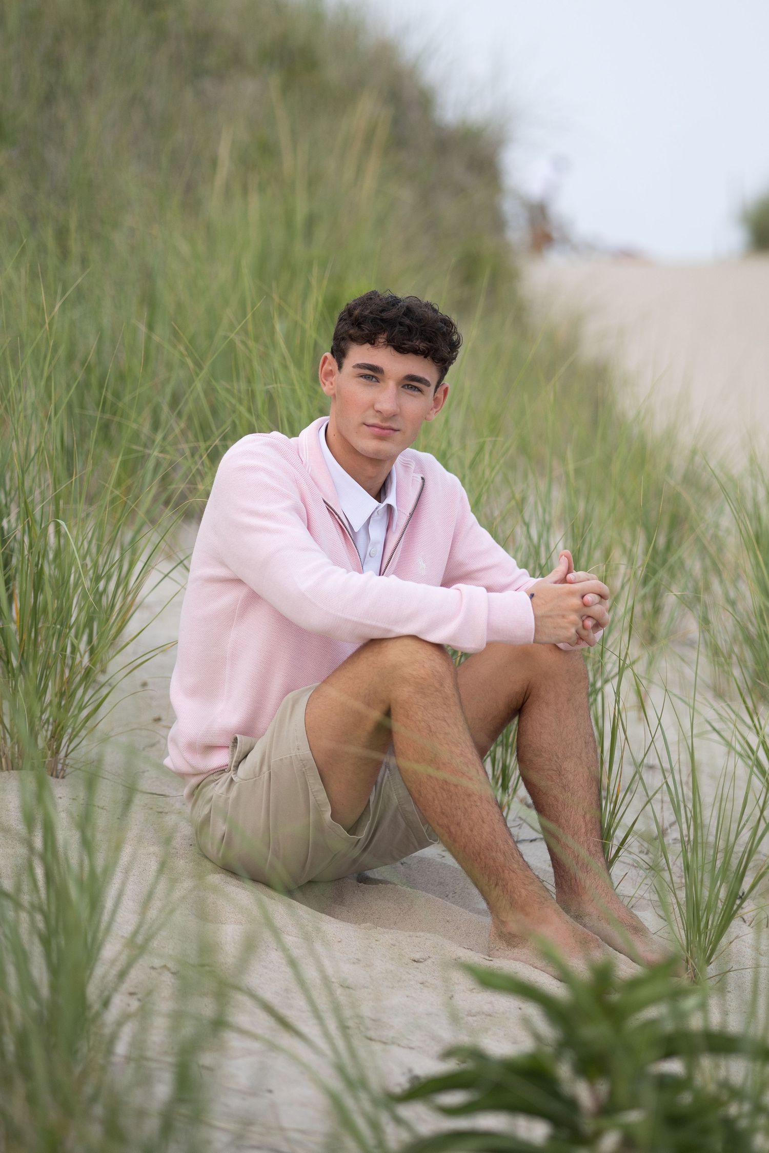 Young person in pink sweater sitting casually on sandy beach dunes surrounded by tall grass.
